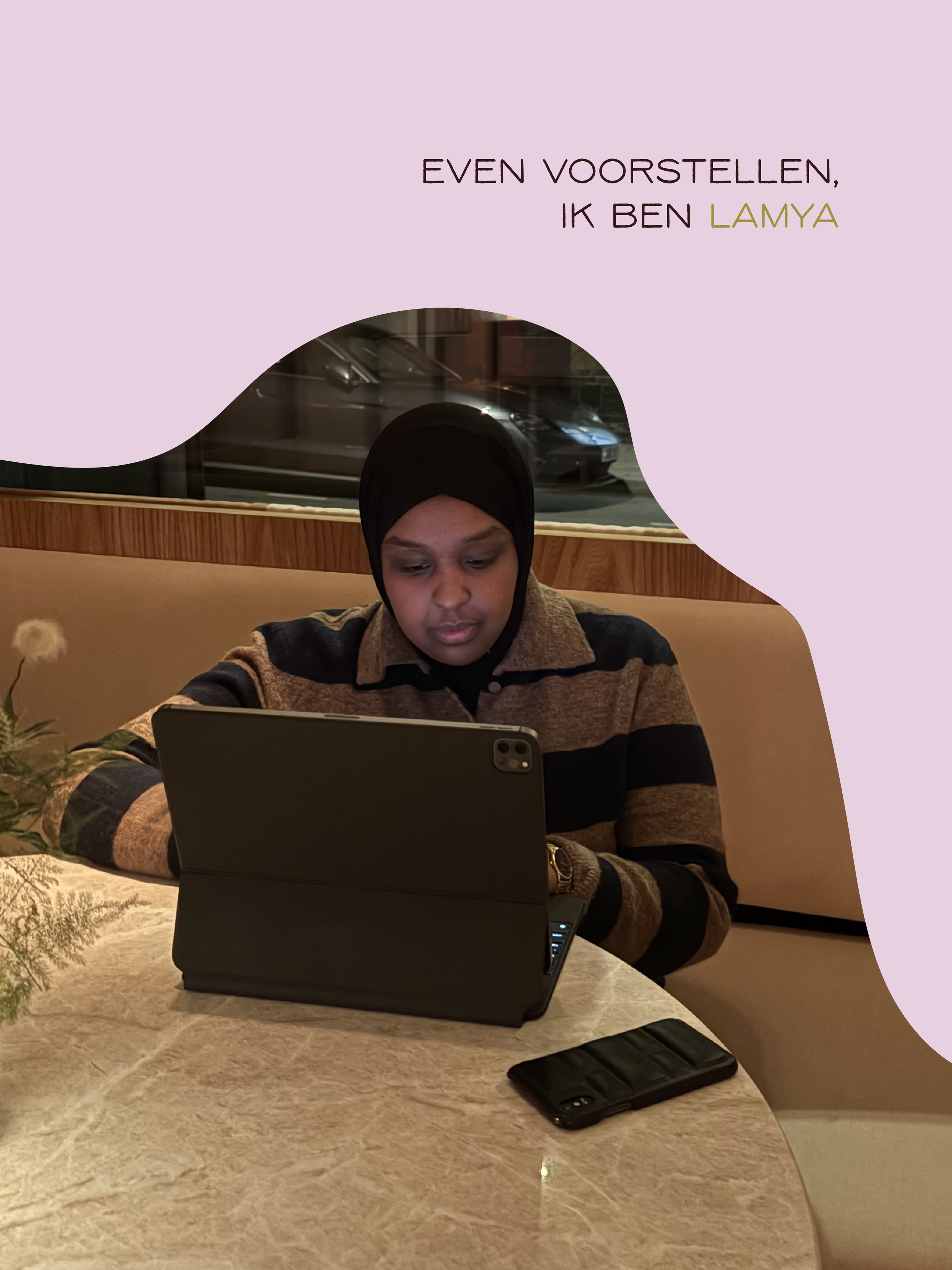 Een vrouw met een hoofddoek werkt achter een laptop aan een tafel in een restaurant of café. Op de tafel ligt ook een mobiele telefoon.