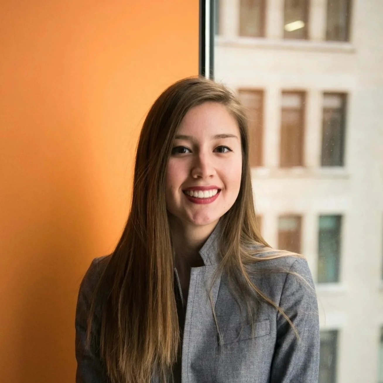 A young woman with long brown hair wearing a gray blazer, smiling, standing by a window with a cityscape background.
