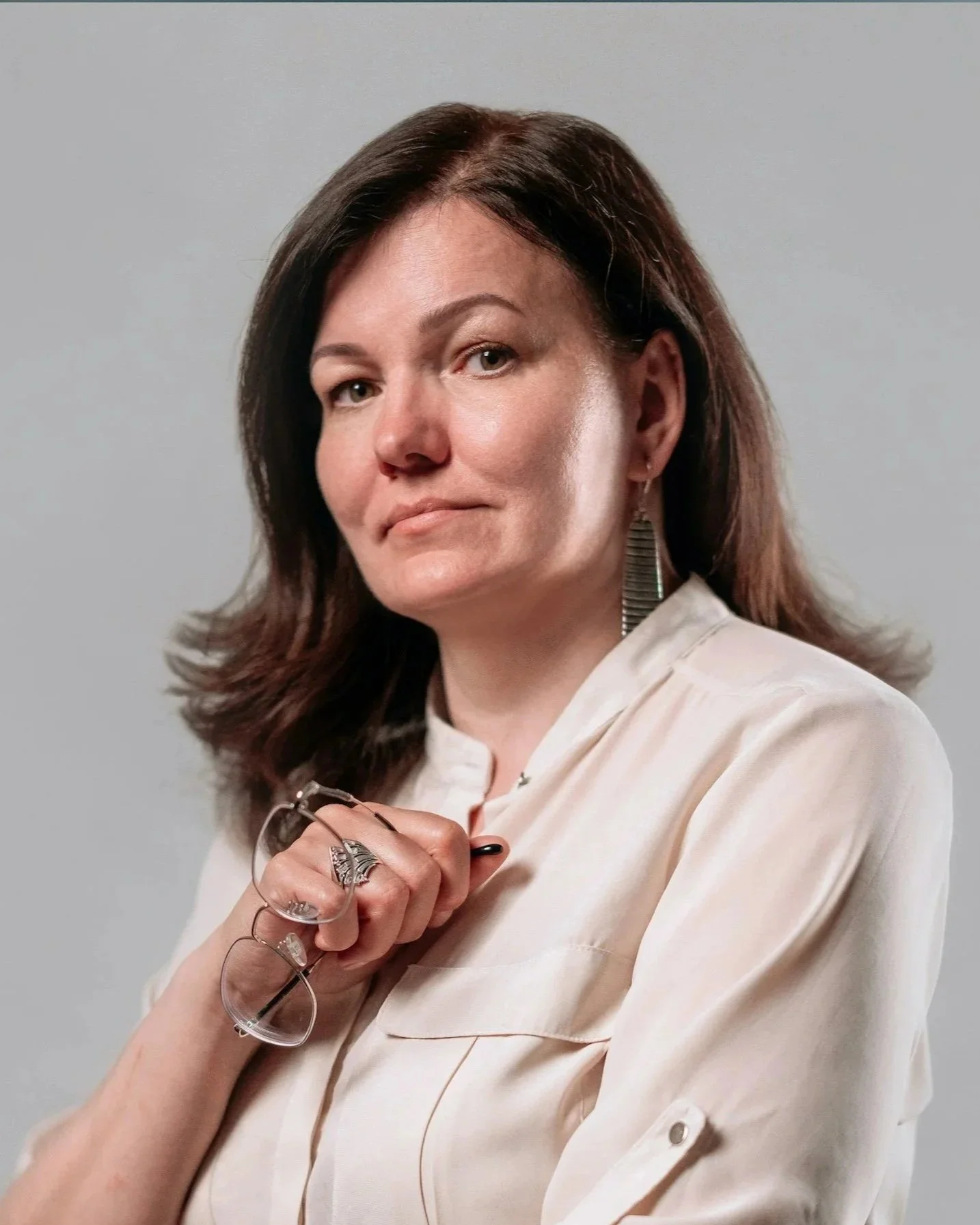 A woman with brown hair, wearing a cream-colored shirt, holding a pair of patterned glasses in front of her chest, looking confidently at the camera against a plain background.