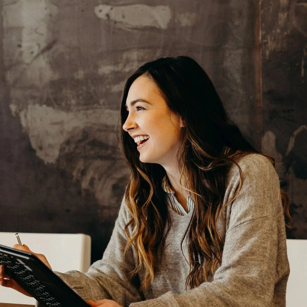 A young woman with long wavy brown hair, wearing a beige sweater, is smiling and looking to her left while sitting at a table, holding a pen and a black folder, with a dark wall in the background.
