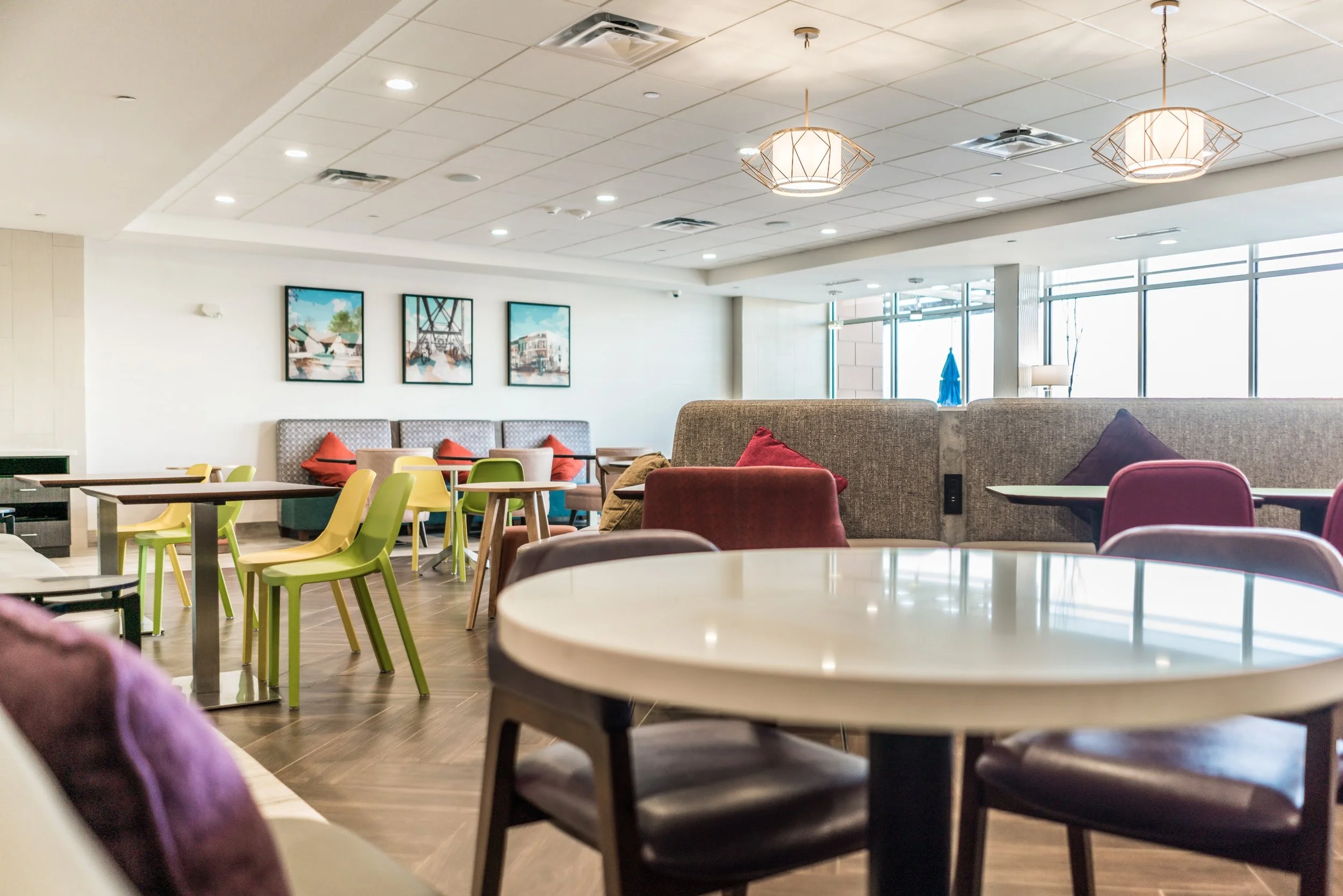 Modern cafeteria with white tables, colorful chairs, and decorative cushions. Framed artwork on walls and pendant lighting on ceiling.