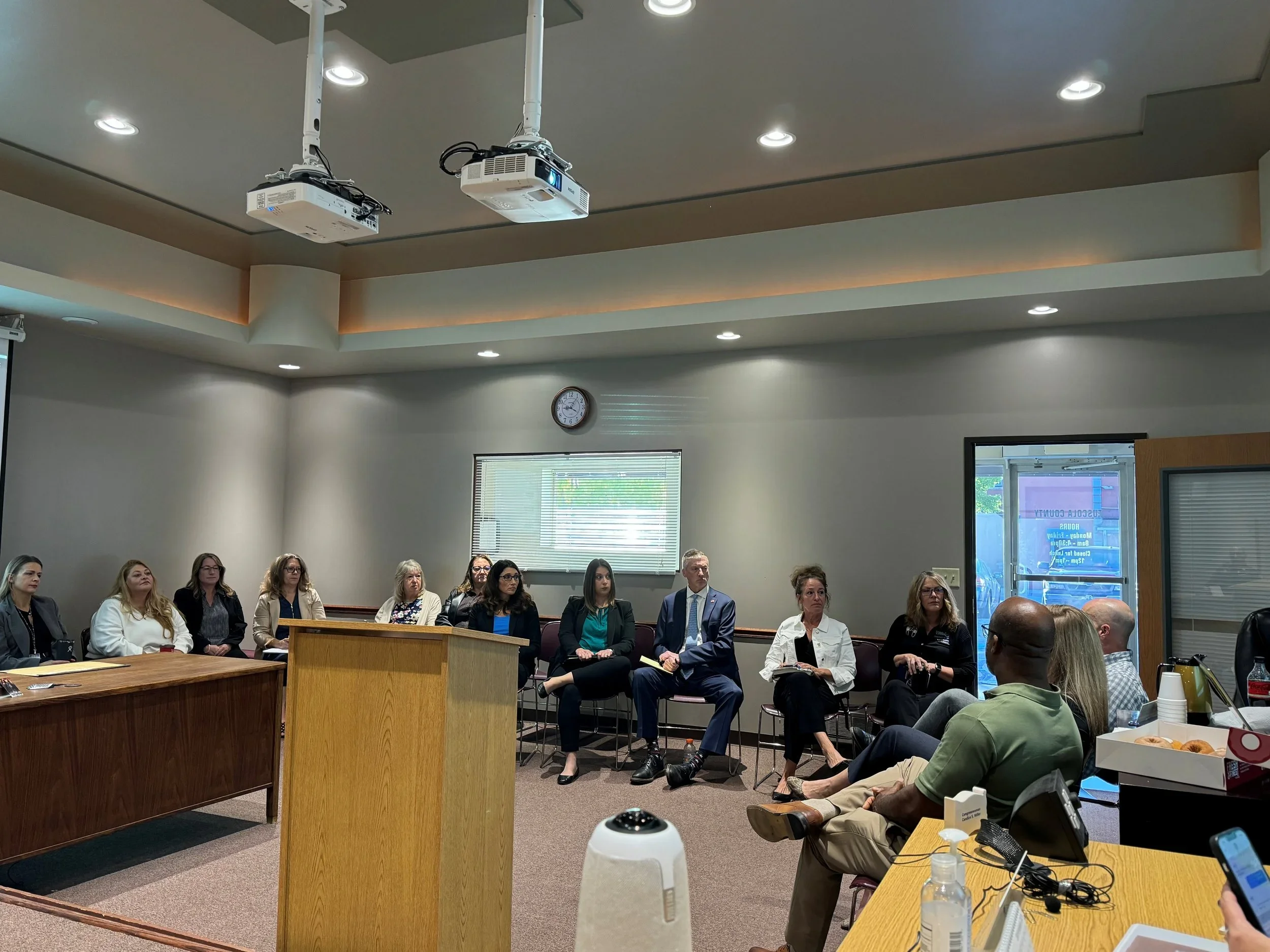A group of people seated in a conference room, listening to a presentation. The room has a ceiling with recessed lighting and two projectors hanging from above. There is a clock on the back wall and a window with blinds. Some people have notebooks, and there is a table with a phone, sanitizer, and snacks in the foreground.