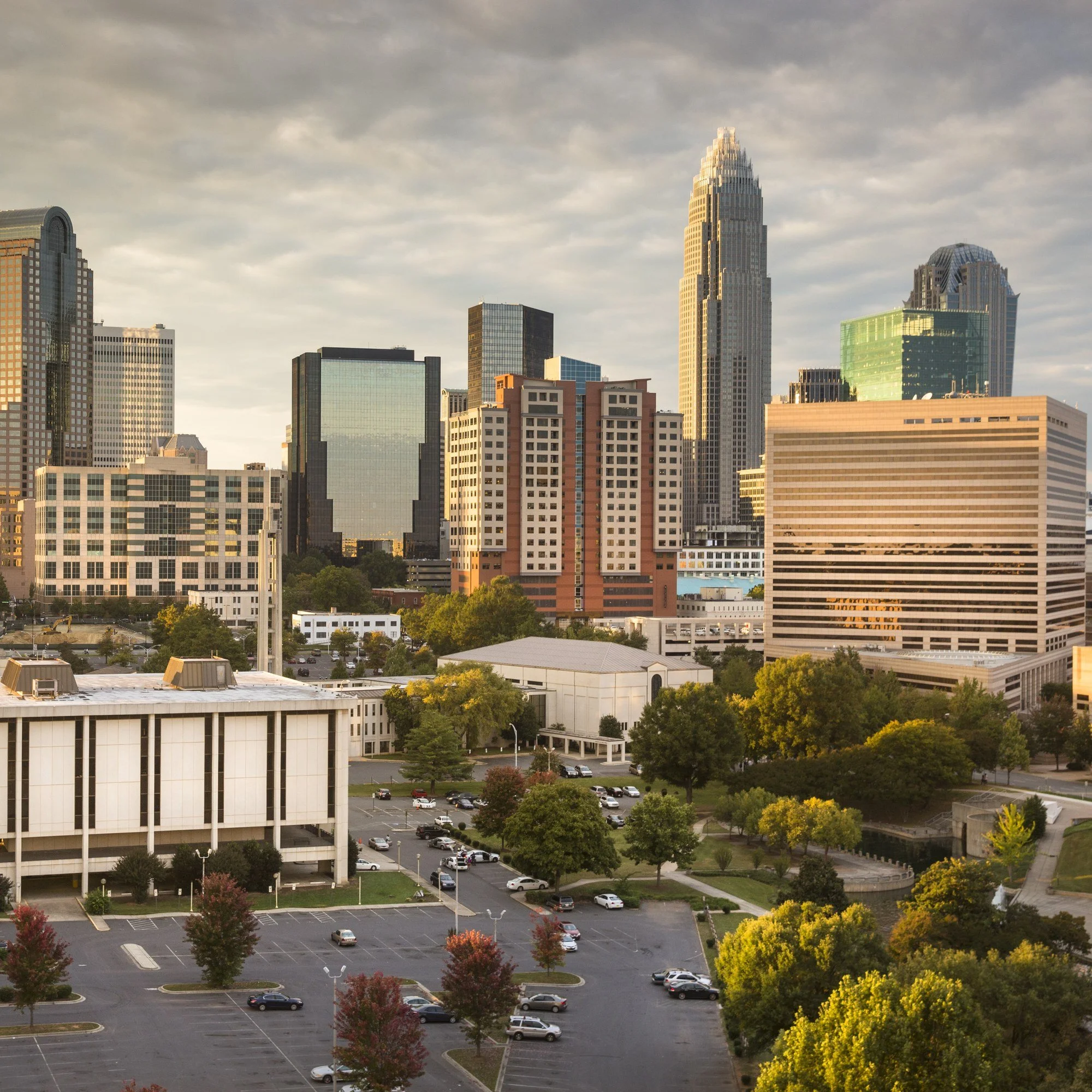 The uptown Charlotte, North Carolina, skyline under heavy clouds, with trees and low buildings in the foreground.