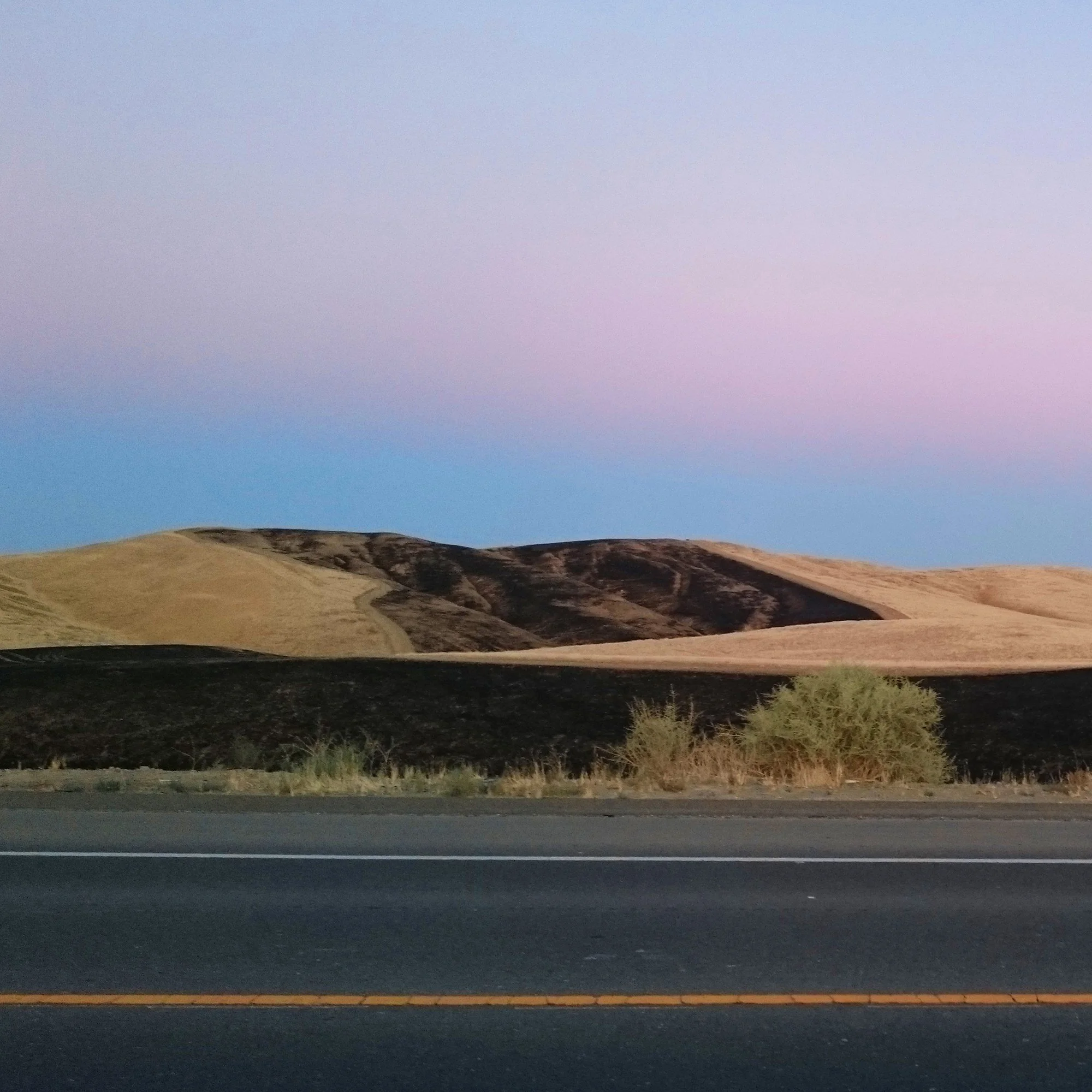 Dry, golden foothills near Kettleman City, California, at dusk, seen from the roadside under a pink and blue sky.