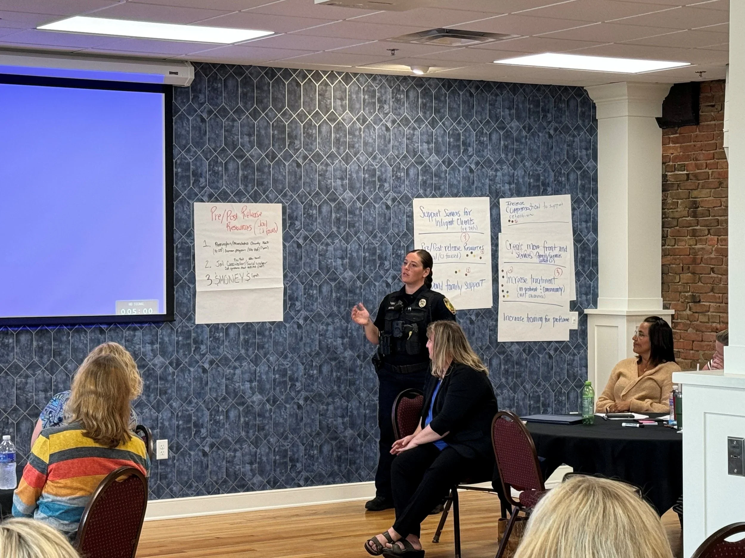 A police officer giving a presentation to a group of women in a conference room. The room has a blue hexagon patterned wall, a large screen with a blue background, and several posters with handwritten notes on the wall.