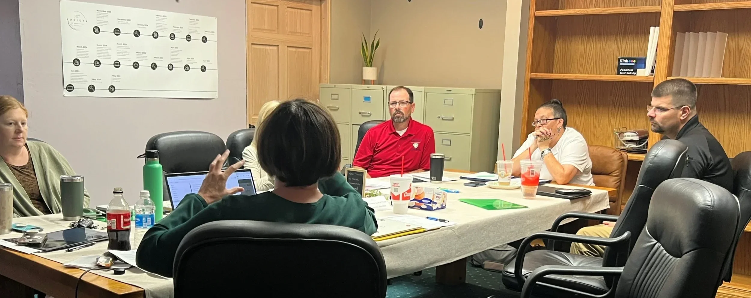 A group of six people sitting around a conference table engaged in a meeting in a room with beige walls and wooden decor. The table has drinks, snacks, laptops, and papers. A large infographic or calendar is on the wall behind them.