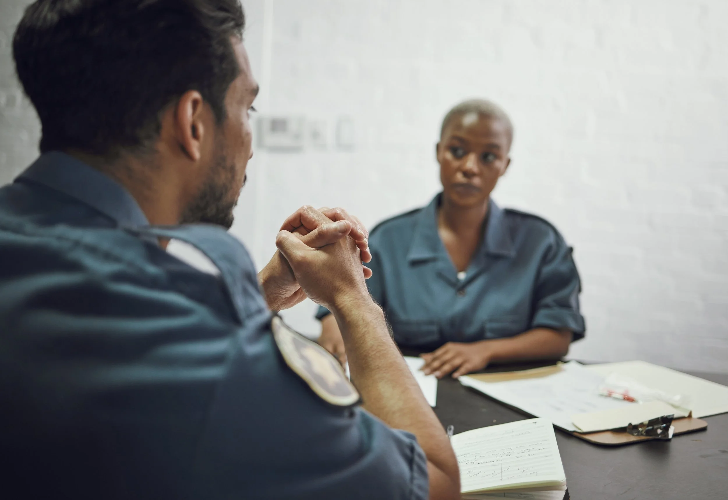 Two uniformed officers sit across a table from each other in conversation, with notes and a clipboard between them.