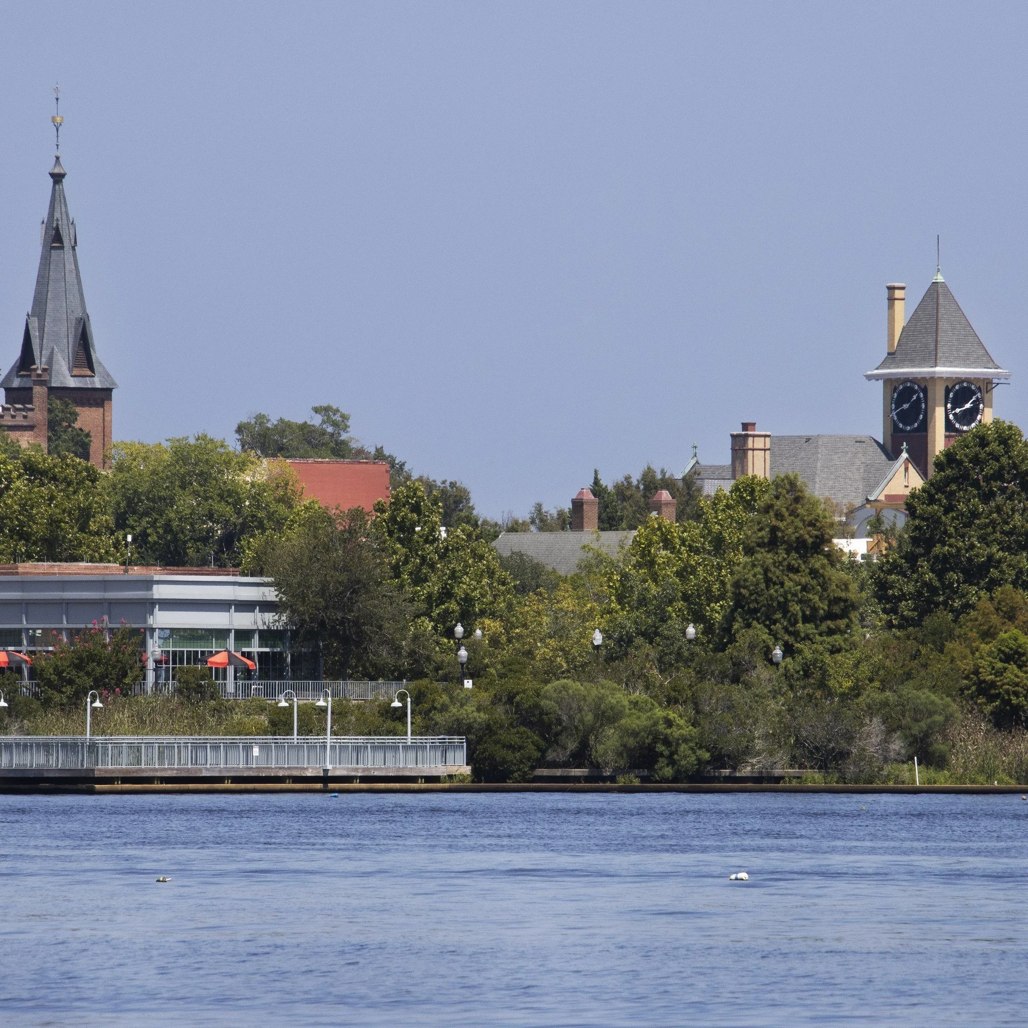 Historic downtown New Bern, North Carolina, seen across the Trent River, with a church steeple and clock tower above the trees.