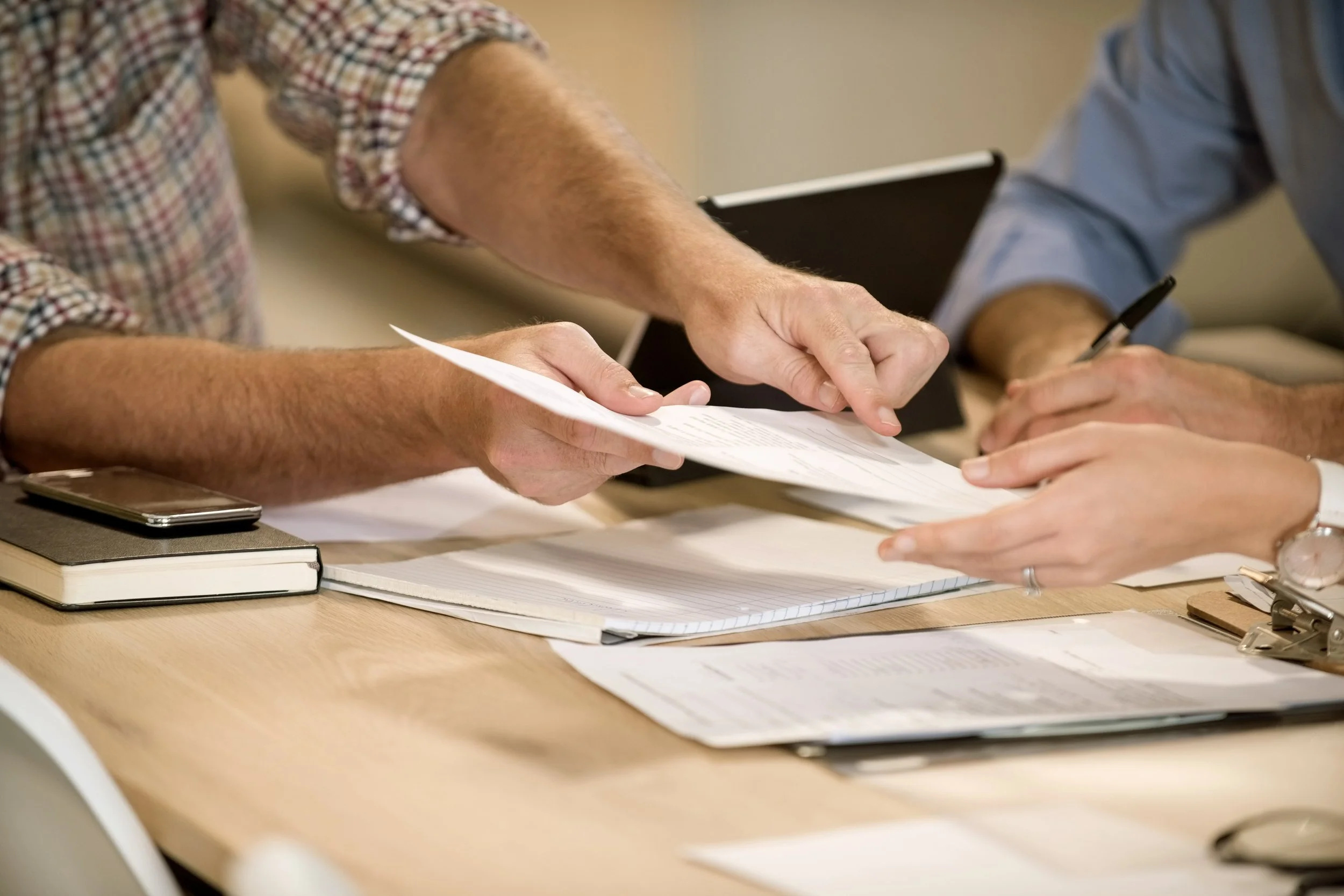 Two people at a desk pass a printed document between them, with notebooks and papers on the table.