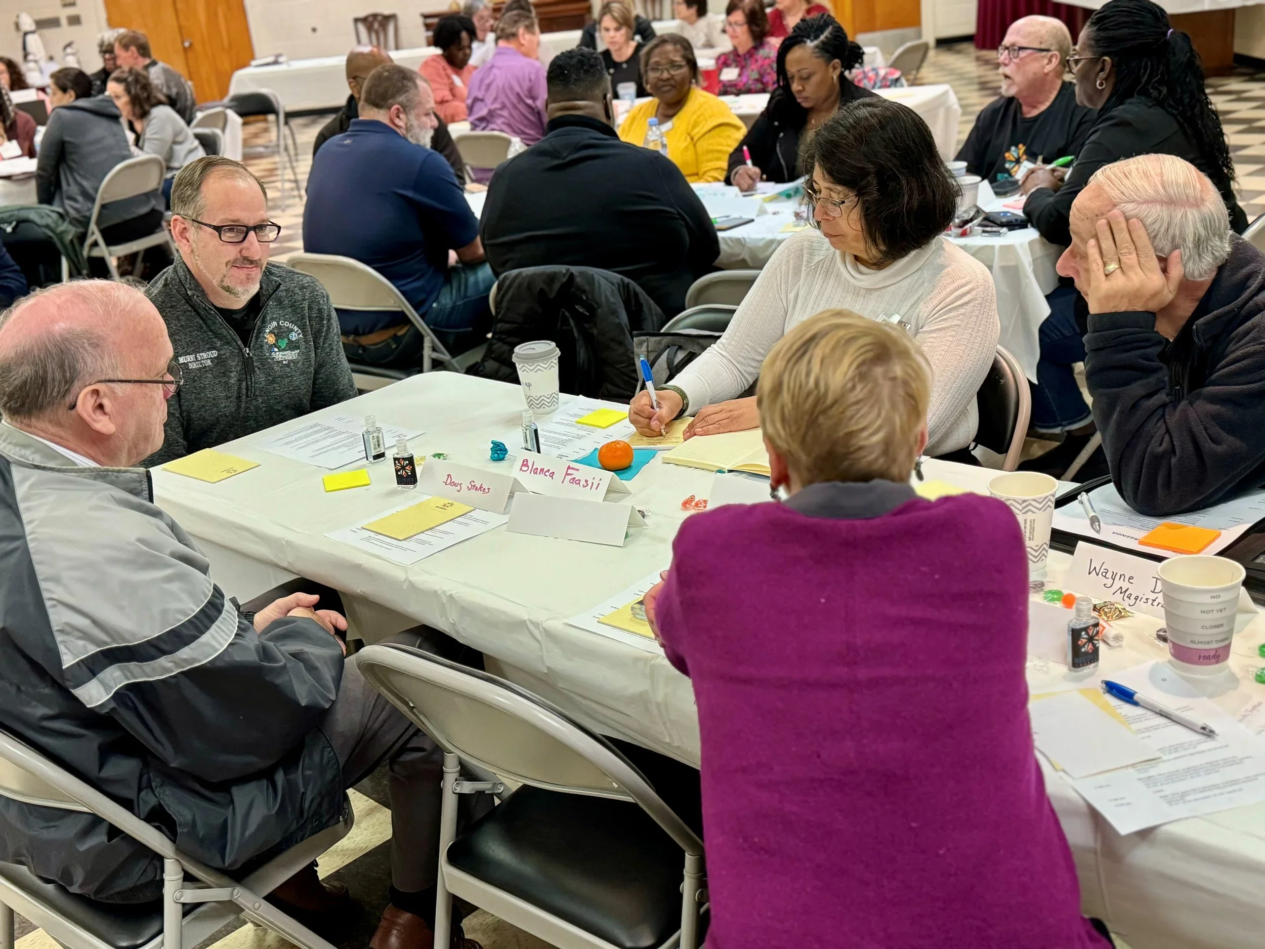 A group of diverse people sitting around a table during a meeting or workshop, some taking notes, in a well-lit conference room with checkered flooring.