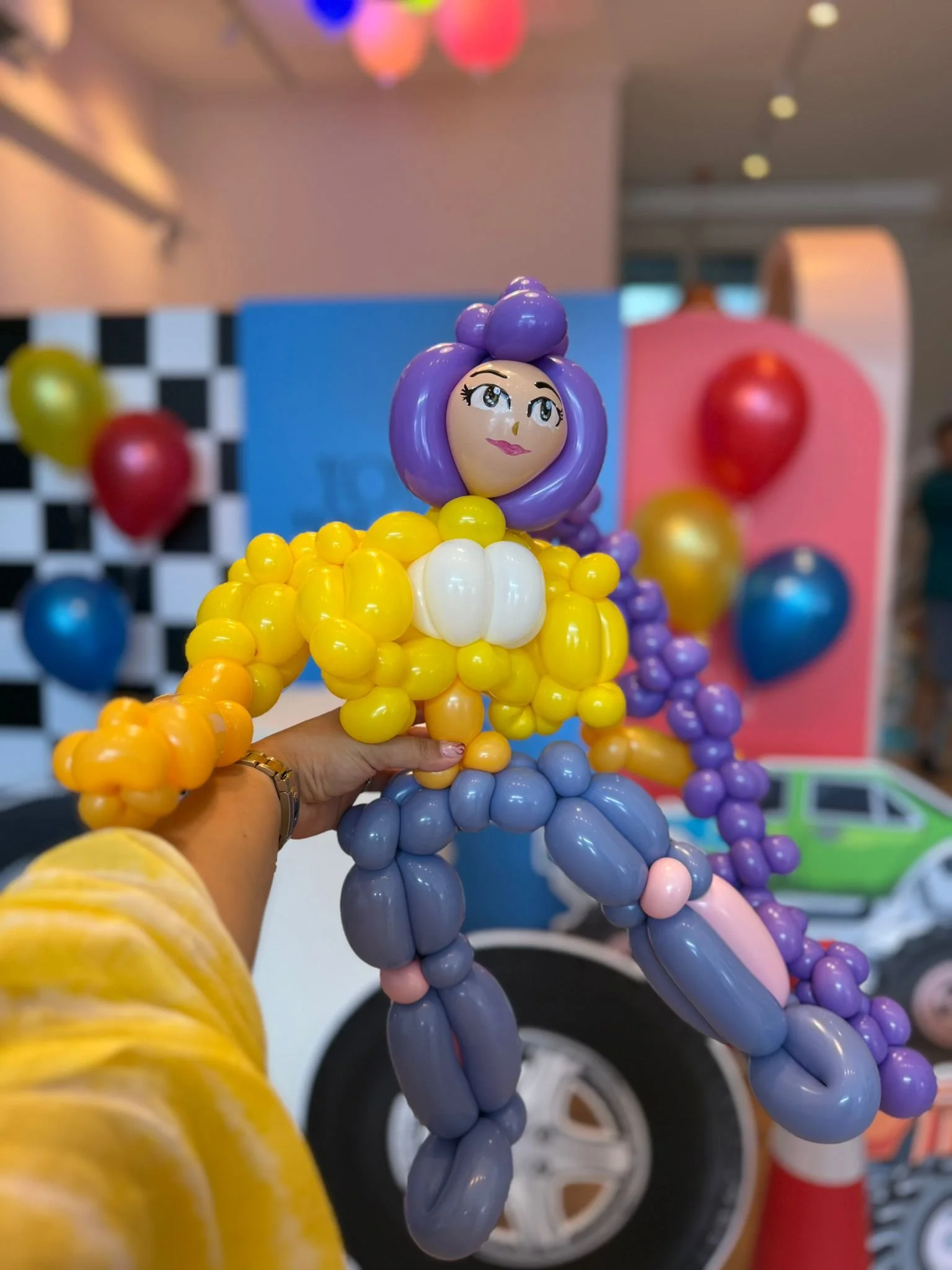 Colorful balloon sculpture resembling a girl with purple hair, a yellow dress, and purple pants, held by a hand at a festive event with balloons and decorations in the background.