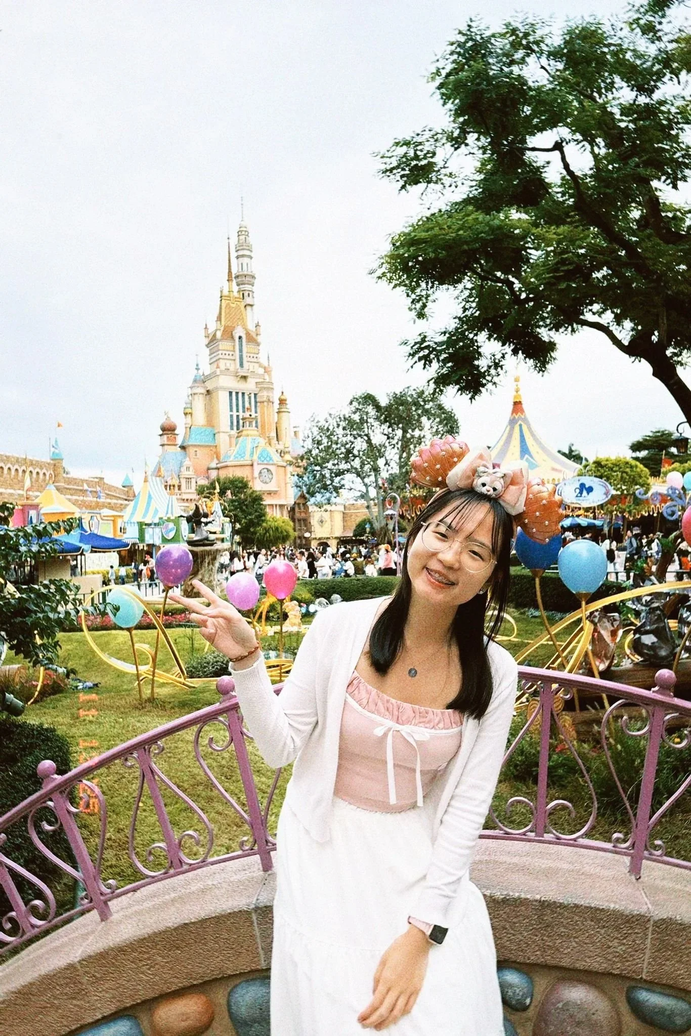 A woman is smiling and posing in front of a Disney castle at a theme park, wearing Mickey Mouse ears and a pastel-colored outfit.