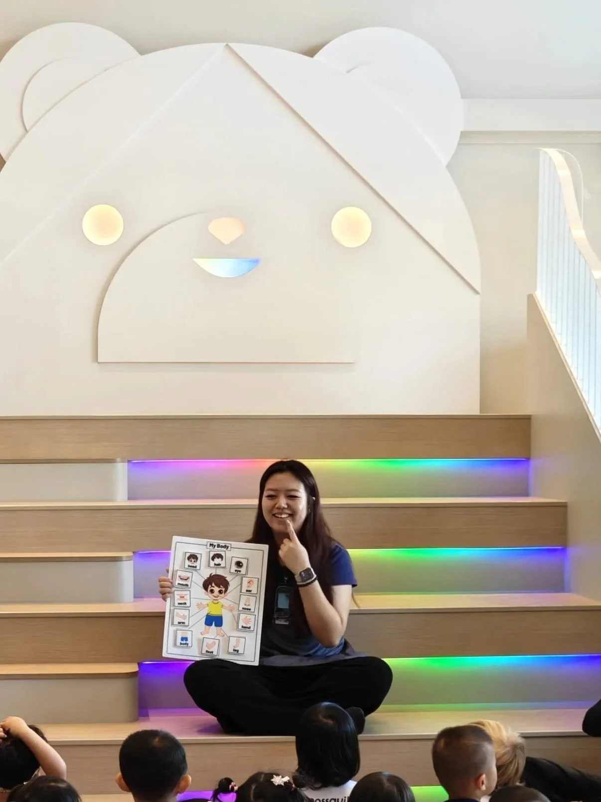A woman sitting on a staircase with rainbow lights, holding a chart about the human body, in front of young children attending an educational session.