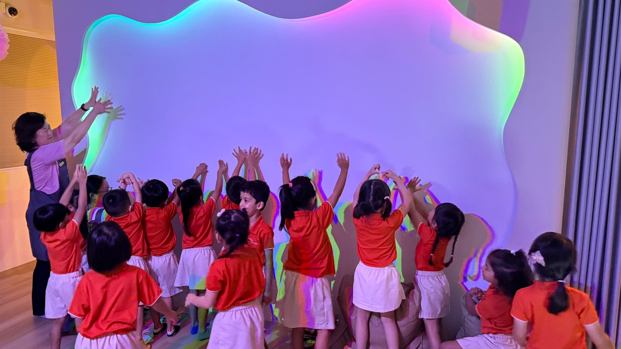 Children in orange uniforms raise their hands towards a wall with colorful light projections, led by a woman in a purple shirt and black apron.