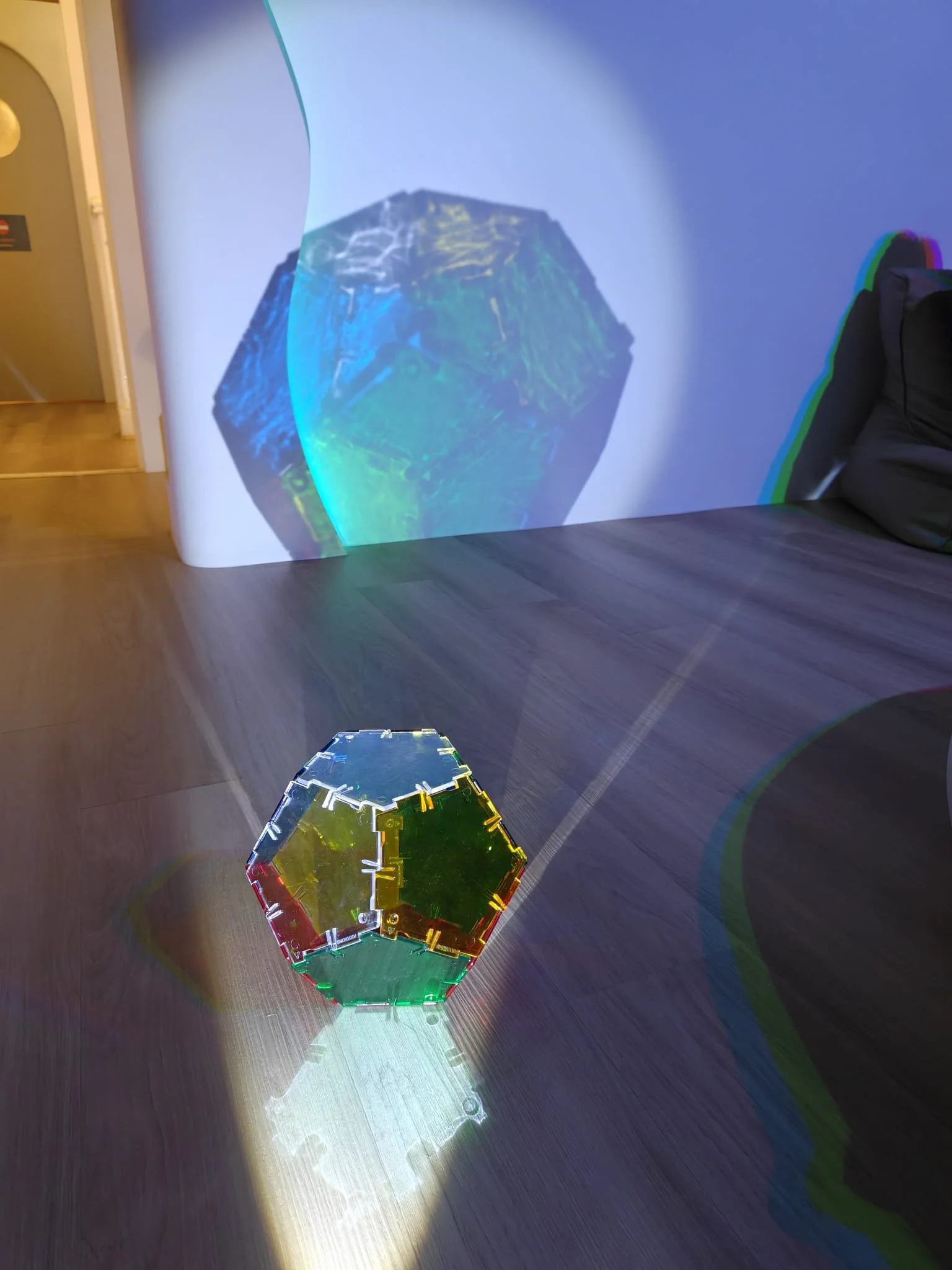 Colorful geometric glass sculpture on a dark wooden floor, casting a colorful shadow on the wall and floor.