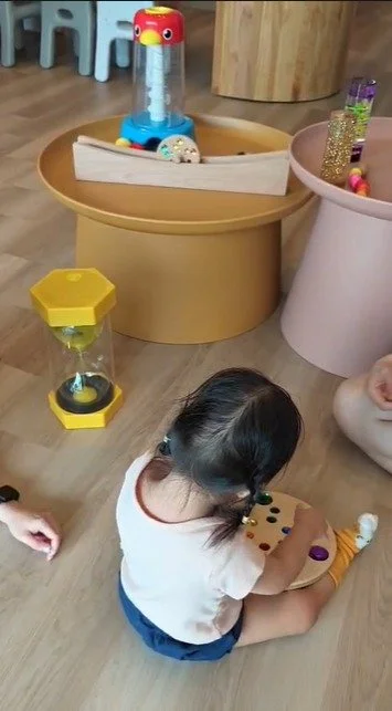 A toddler sitting on the floor playing with a wooden toy. Behind the child, there are decorative items including a yellow and clear hourglass-shaped timer, a wooden boat, and a pink container filled with shiny objects.