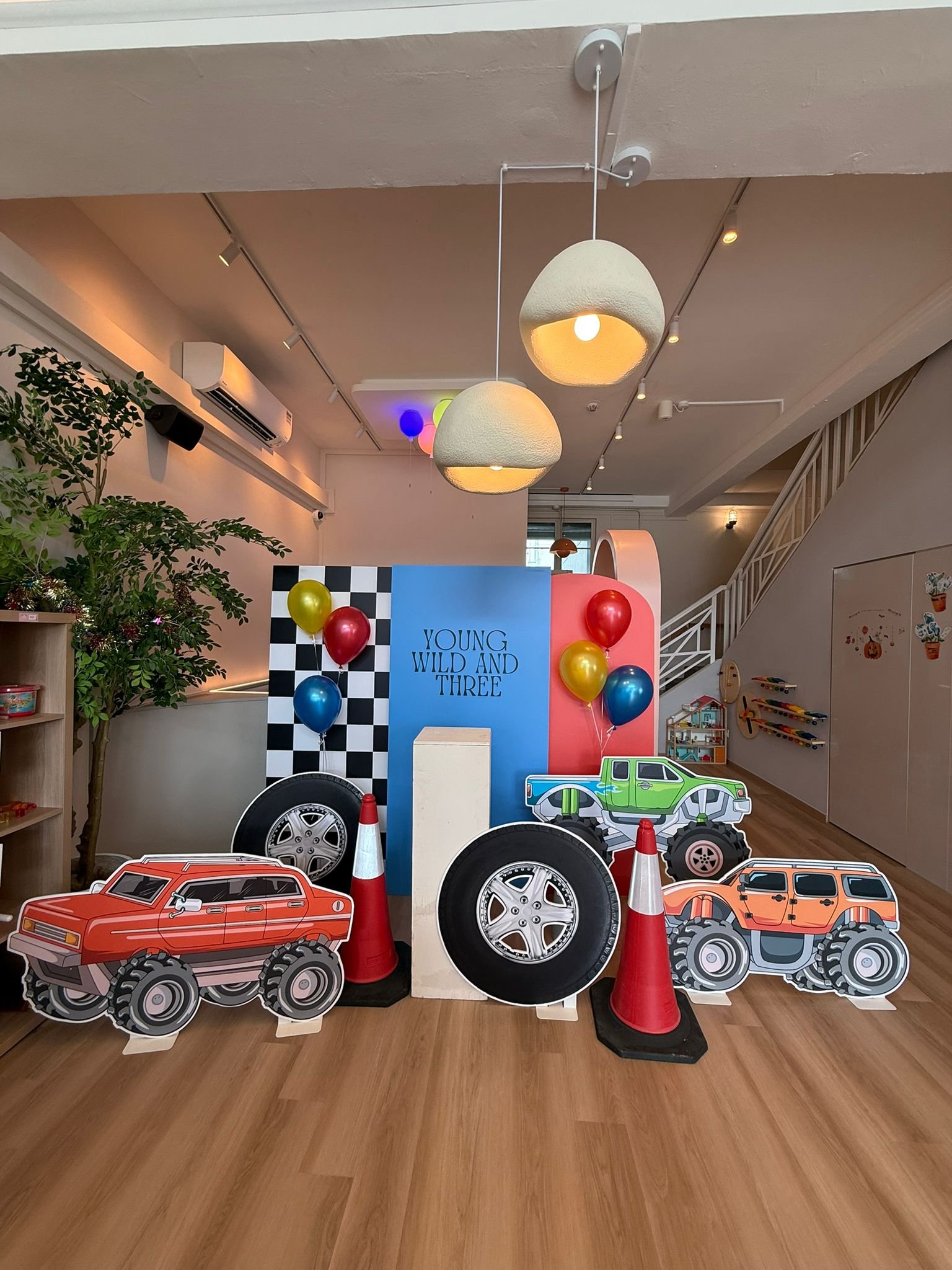 Indoor display with cardboard monster trucks, colorful balloons, and traffic cones, set against a backdrop with the phrase 'YOUNG WILD AND THREE' and checkered pattern.