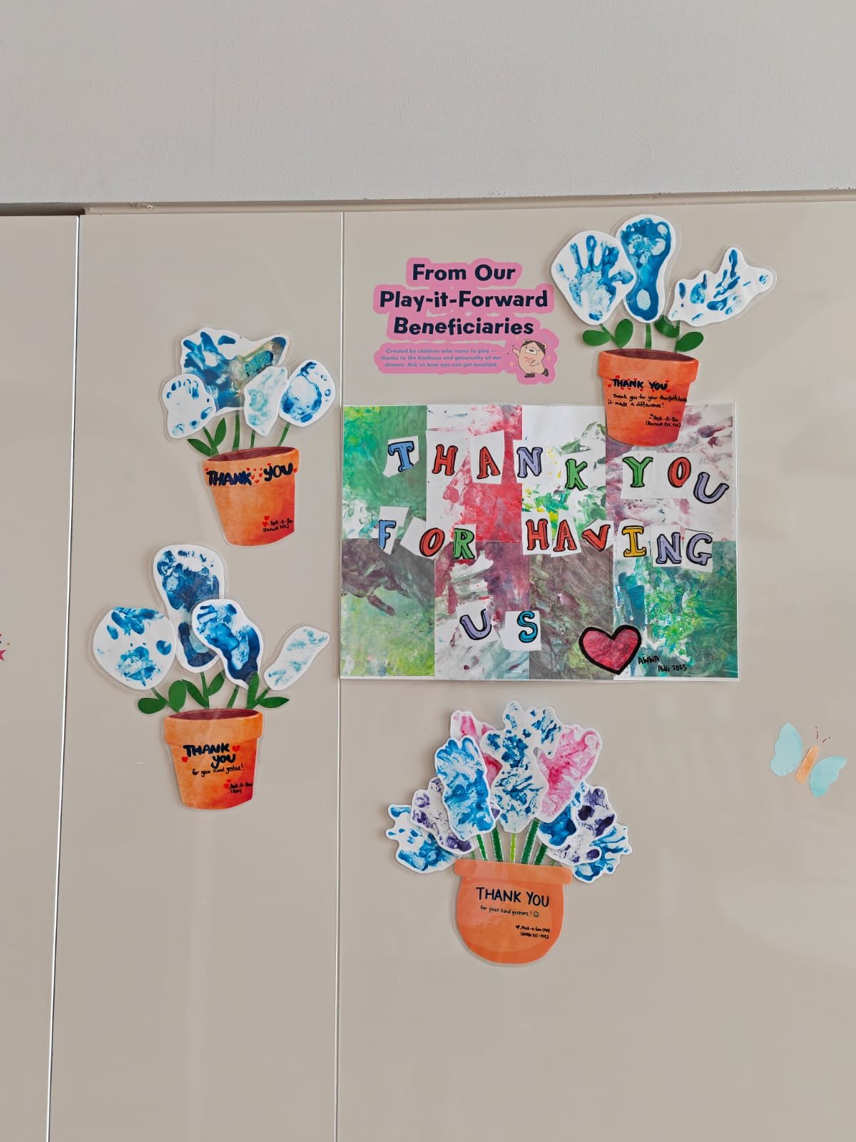 Decorative school bulletin board with paper flowers in pots, colorful cut-out letters spelling "Thank You for Having Us," and a pink sign thanking beneficiaries, labeled "From Our Play-it-Forward Beneficiaries."