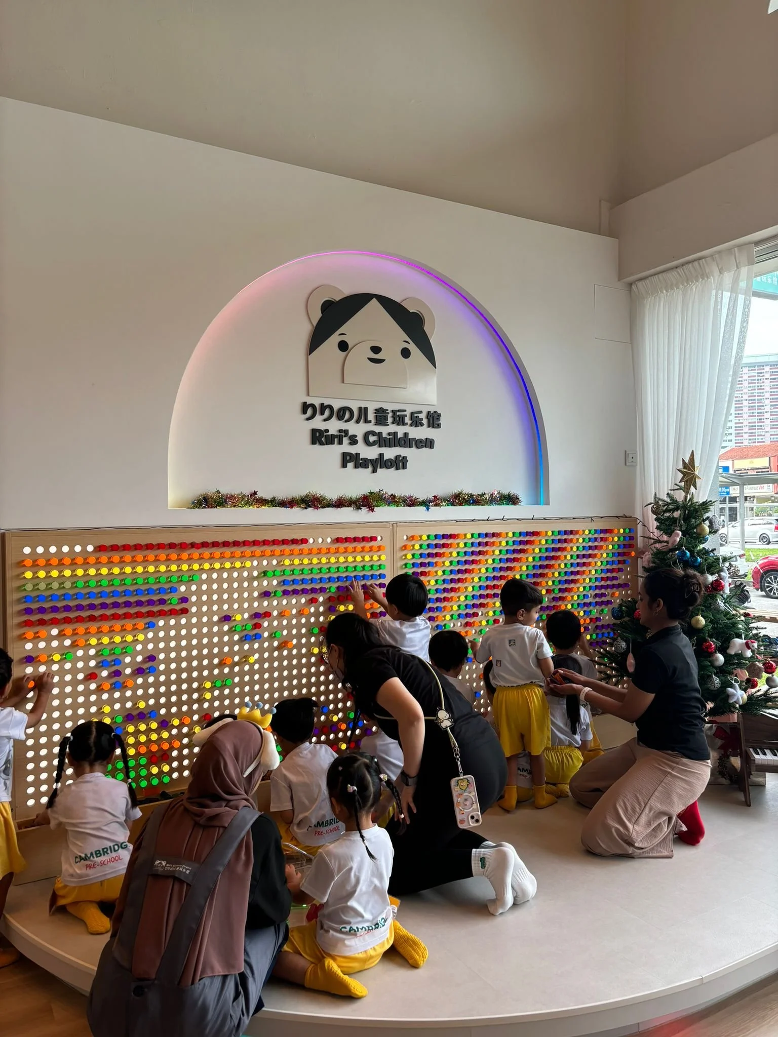 Children playing with a light-up pegboard in a children's play area, with a Christmas tree nearby and a sign that reads 'Riri's Children Playoff' on the wall.