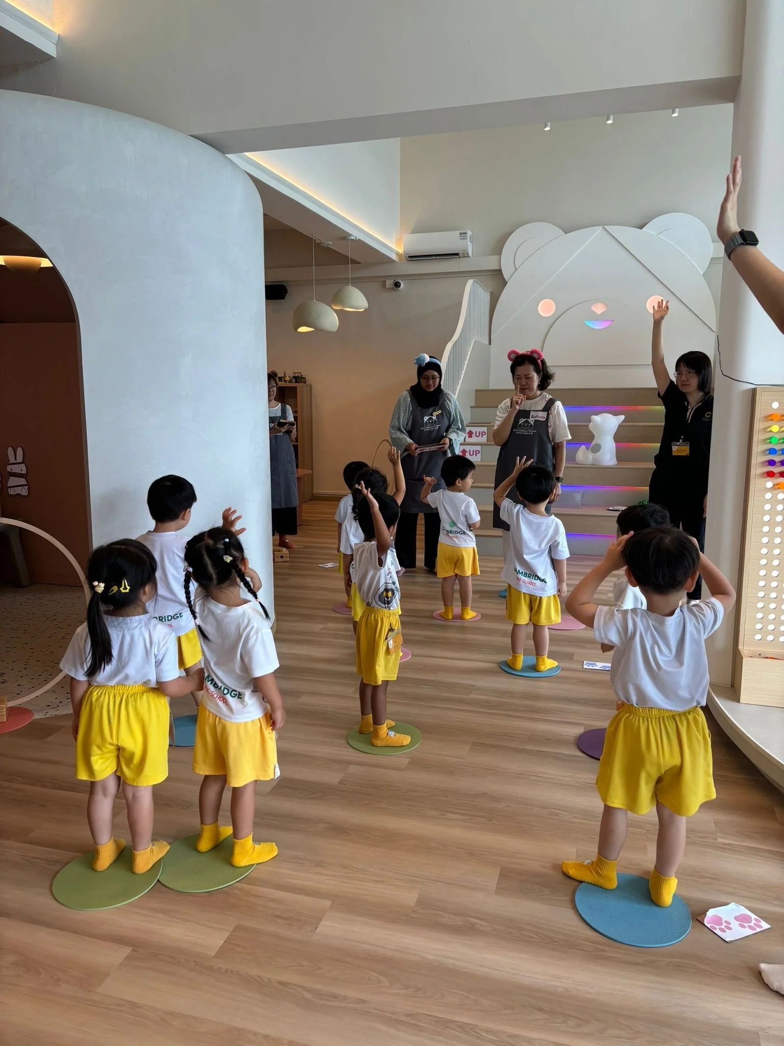 Children in yellow shorts and white shirts participating in an indoor activity, standing on colorful circular mats, with three adults leading the activity near a staircase with a bear face design.