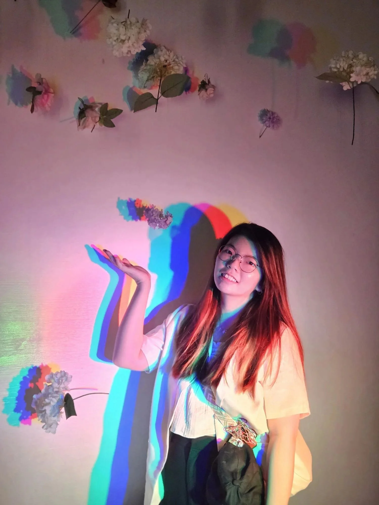 A woman with long reddish hair and glasses smiling at the camera, standing against a wall with floral decorations and colorful shadows cast by a multicolored light.