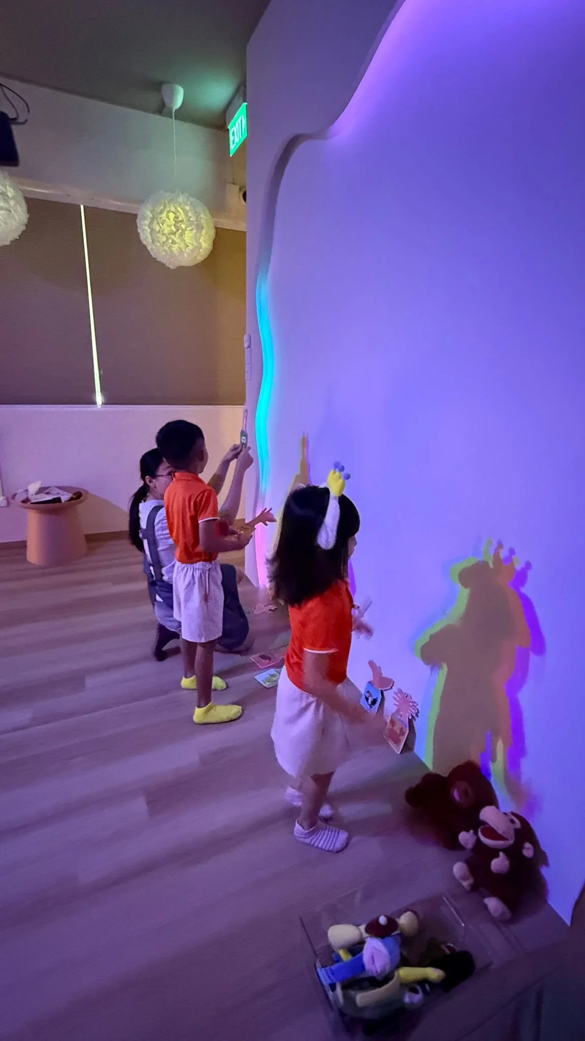 Children engaging with a colorful, interactive wall display in a dimly lit room, with plush toys on the floor nearby.
