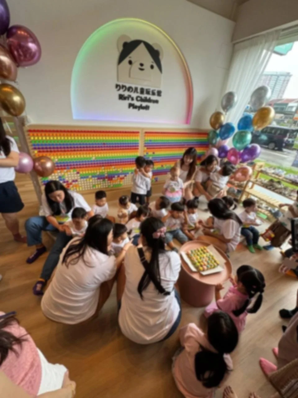 Group of children and adults sitting and standing around a table with a cake, inside a decorated room with balloons and a large, colorful wall art display with a logo reading 'Reina's Culture Played' and a cartoon character of a girl with black hair 