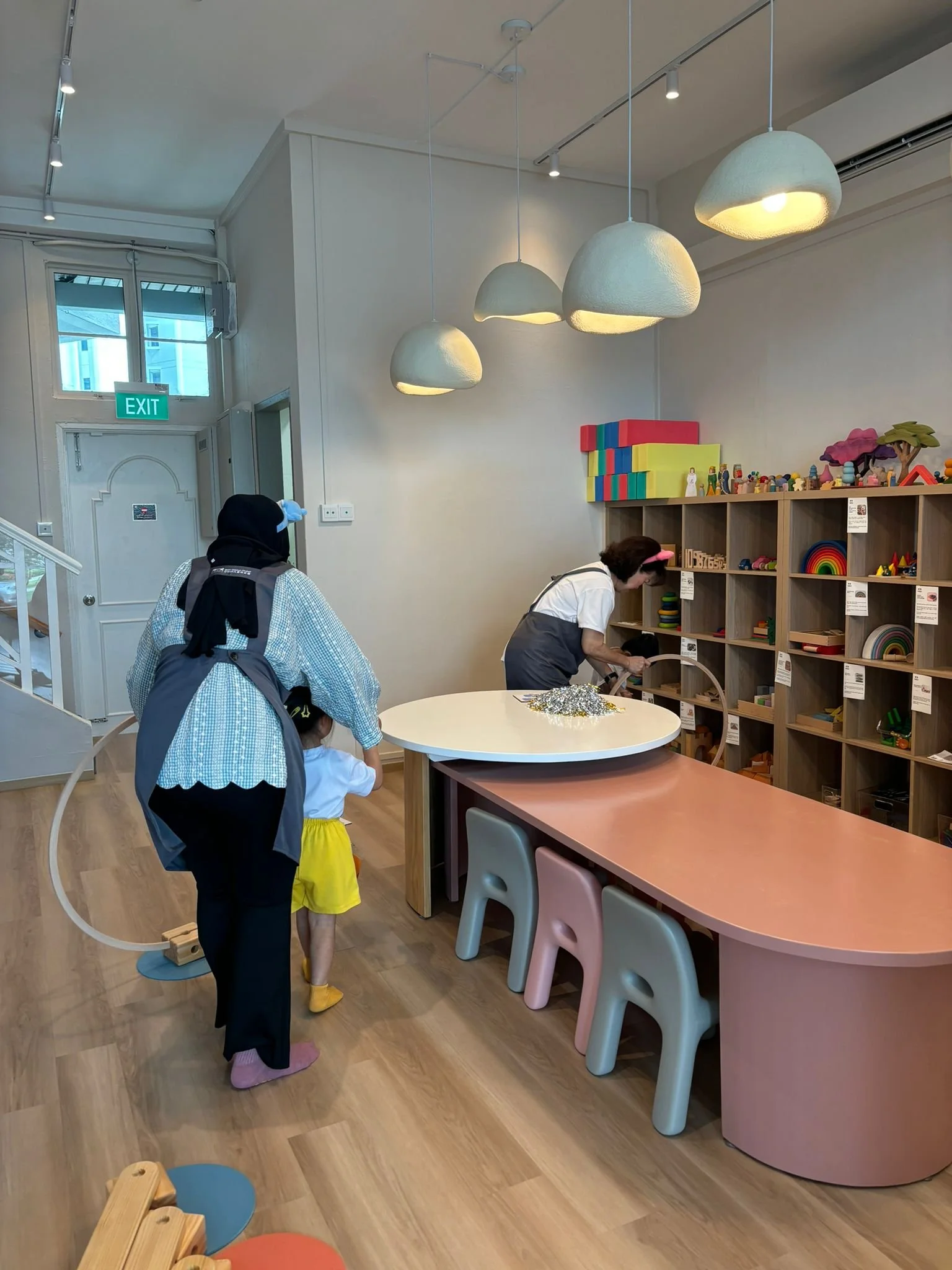 A woman and a girl with a woman in a white shirt and gray apron standing near a wooden shelf with colorful toys and educational materials in a bright, modern playroom. The woman and girl are looking at the toys, and the woman is bending over to exami