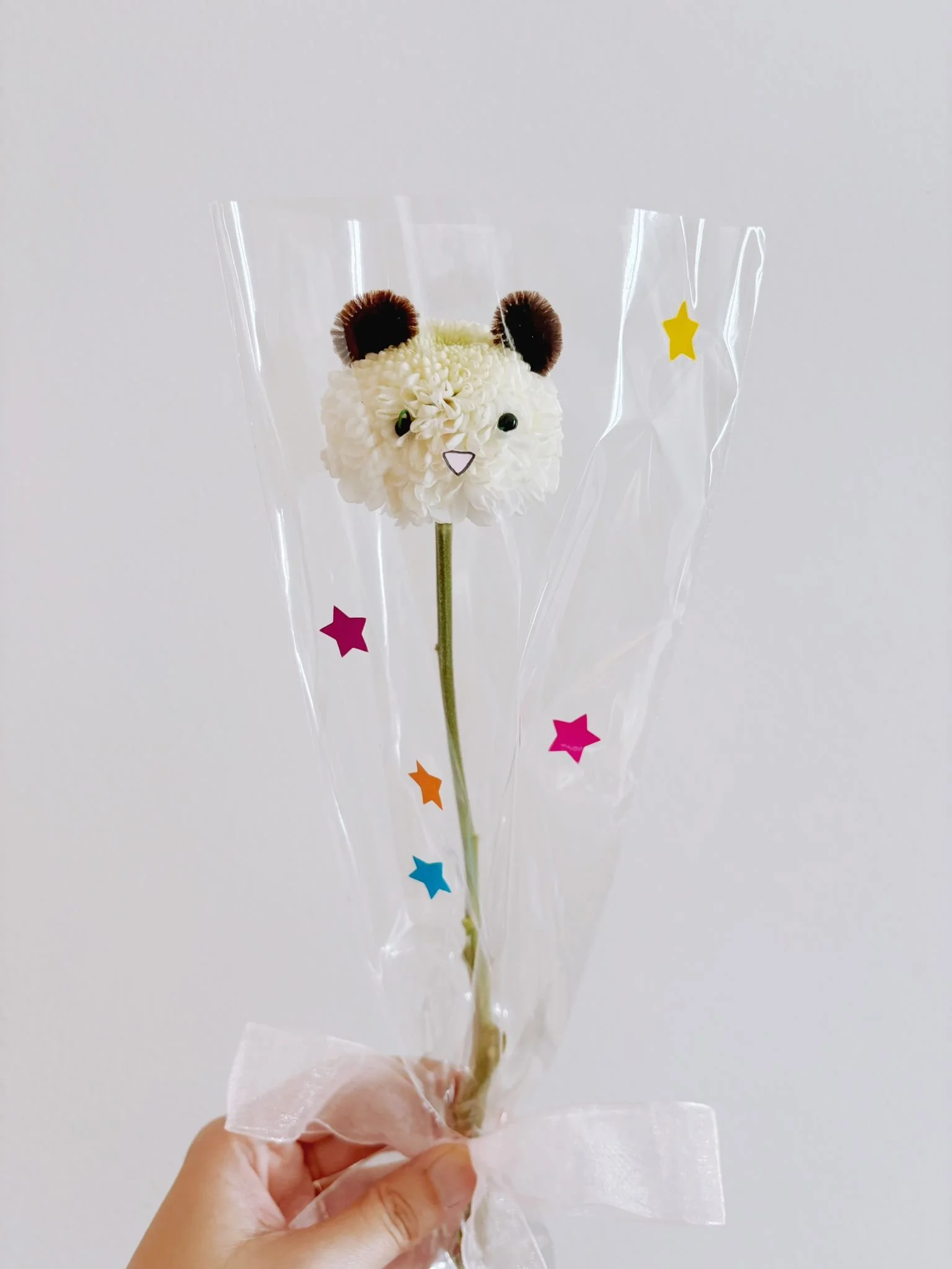 A flower with a bear face on top, wrapped in clear plastic with colorful star-shaped stickers, and tied with a pink ribbon.