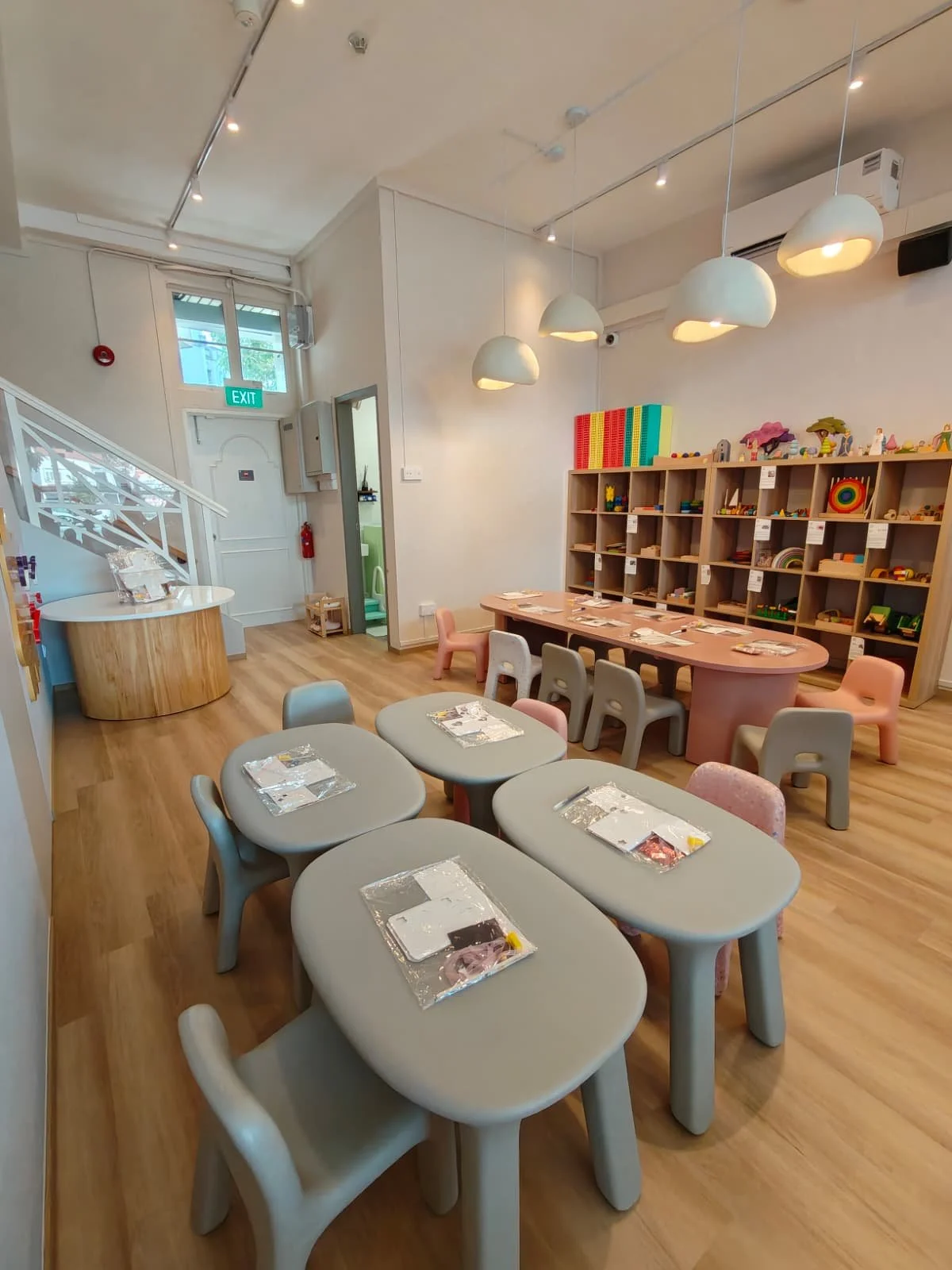 A children's play area with small tables and chairs, wooden shelves filled with toys, and a staircase in the background.