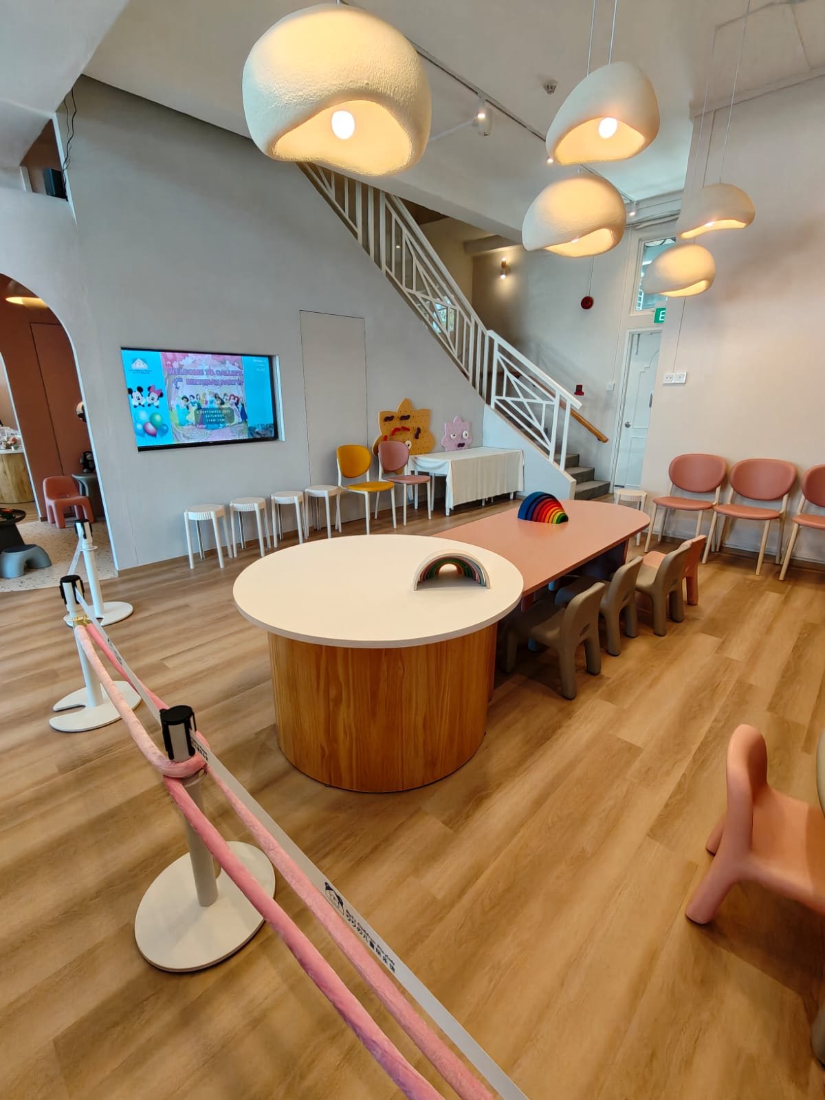 Indoor children's play area with small chairs, tables, stuffed animal decor, and colorful toys. Modern lighting fixtures hang from the ceiling, and a staircase leads to an upper level.
