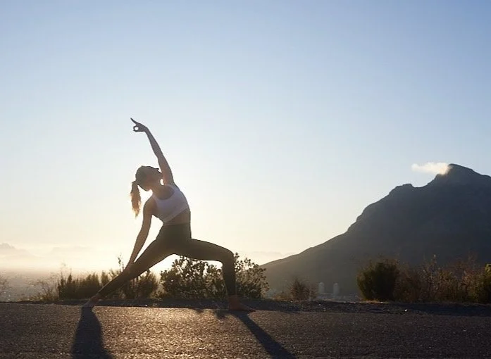 Frau in Yogapose im Freien bei Sonnenuntergang, Berge im Hintergrund.