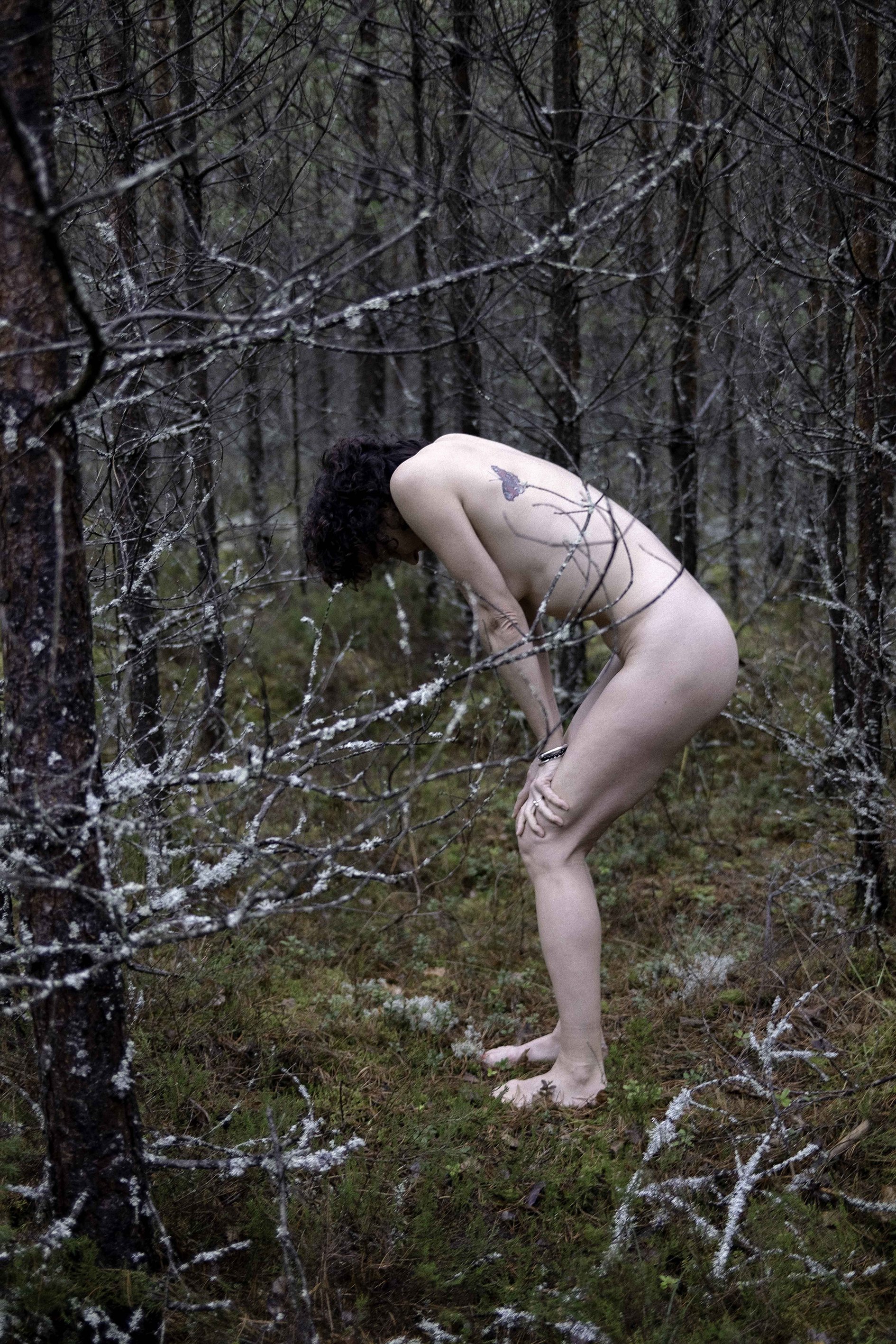 Nude person standing in a forest surrounded by trees and moss.
