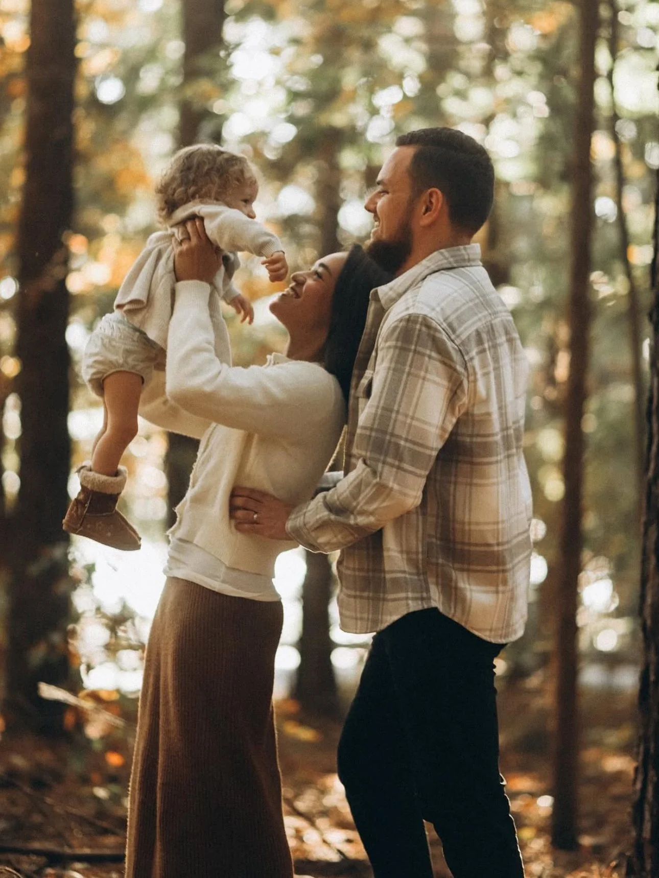 A family enjoying a moment in a wooded area, with a woman holding a baby and a man standing beside her.