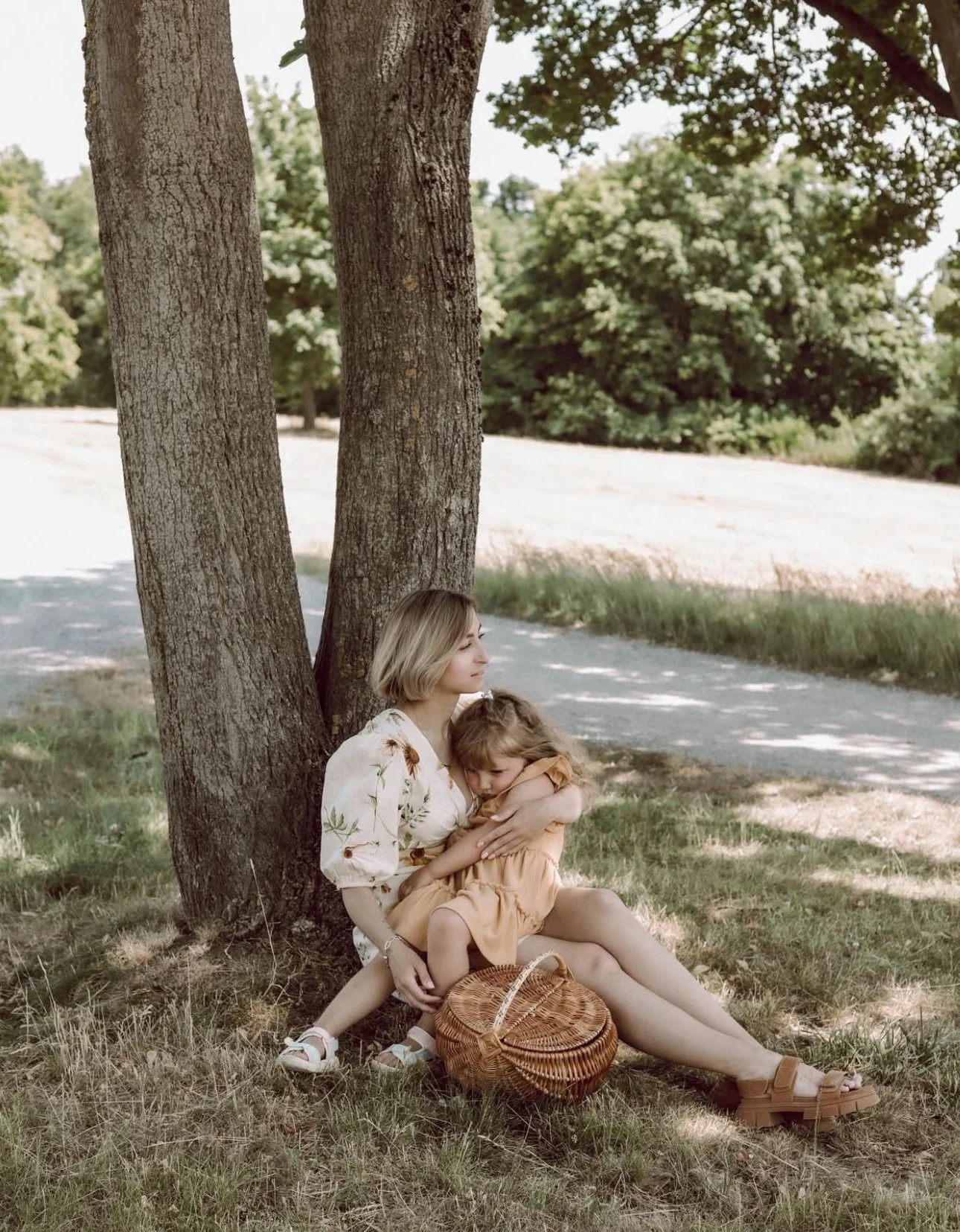 A woman and a child sitting under a tree, embracing each other, with a wicker basket nearby in a sunny, grassy area.