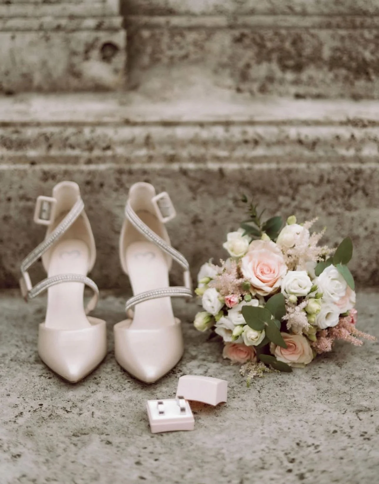 Elegant strappy high heels next to a bouquet of pink and white flowers and a small jewelry box on a textured stone background.