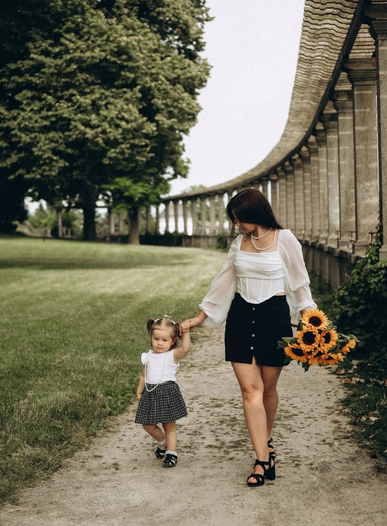 A woman holding a bouquet of sunflowers and the hand of a young girl. They are walking on a path next to a hedge and an arched stone structure. The woman is wearing a white blouse and a black skirt, while the girl is in a white top and plaid skirt.