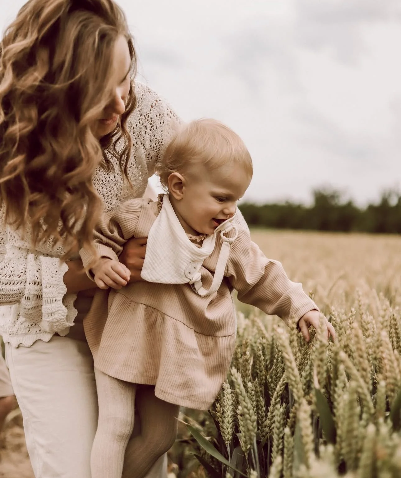 Woman holding a toddler in a wheat field, both wearing beige clothing.