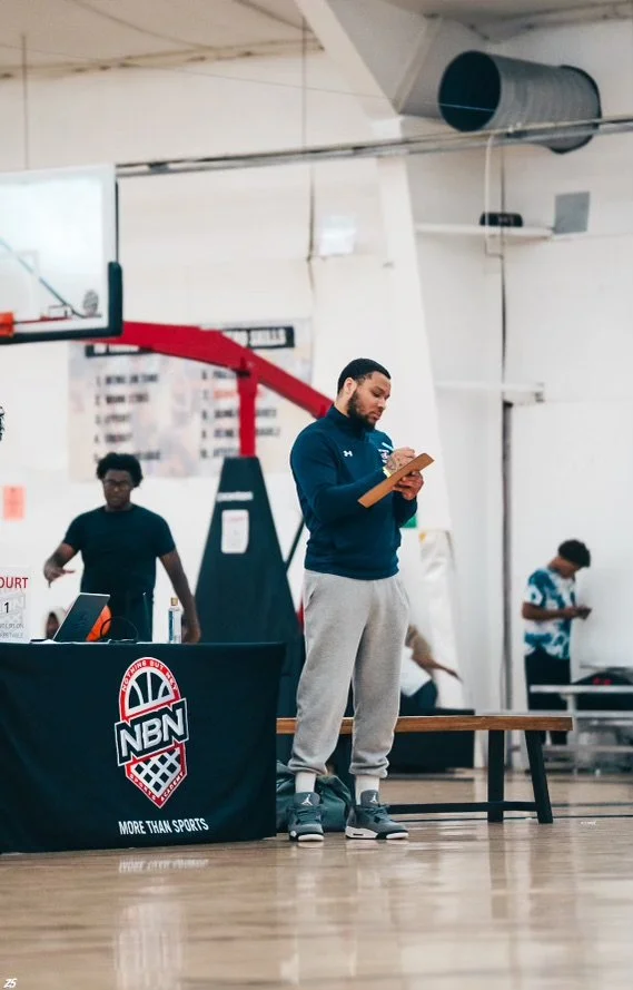 A coach standing on a basketball court with a notepad during a basketball practice or game. There are two players in the background and a table with a logo reading "NBN" and "More Than Sports." The setting appears to be an indoor gymnasium.