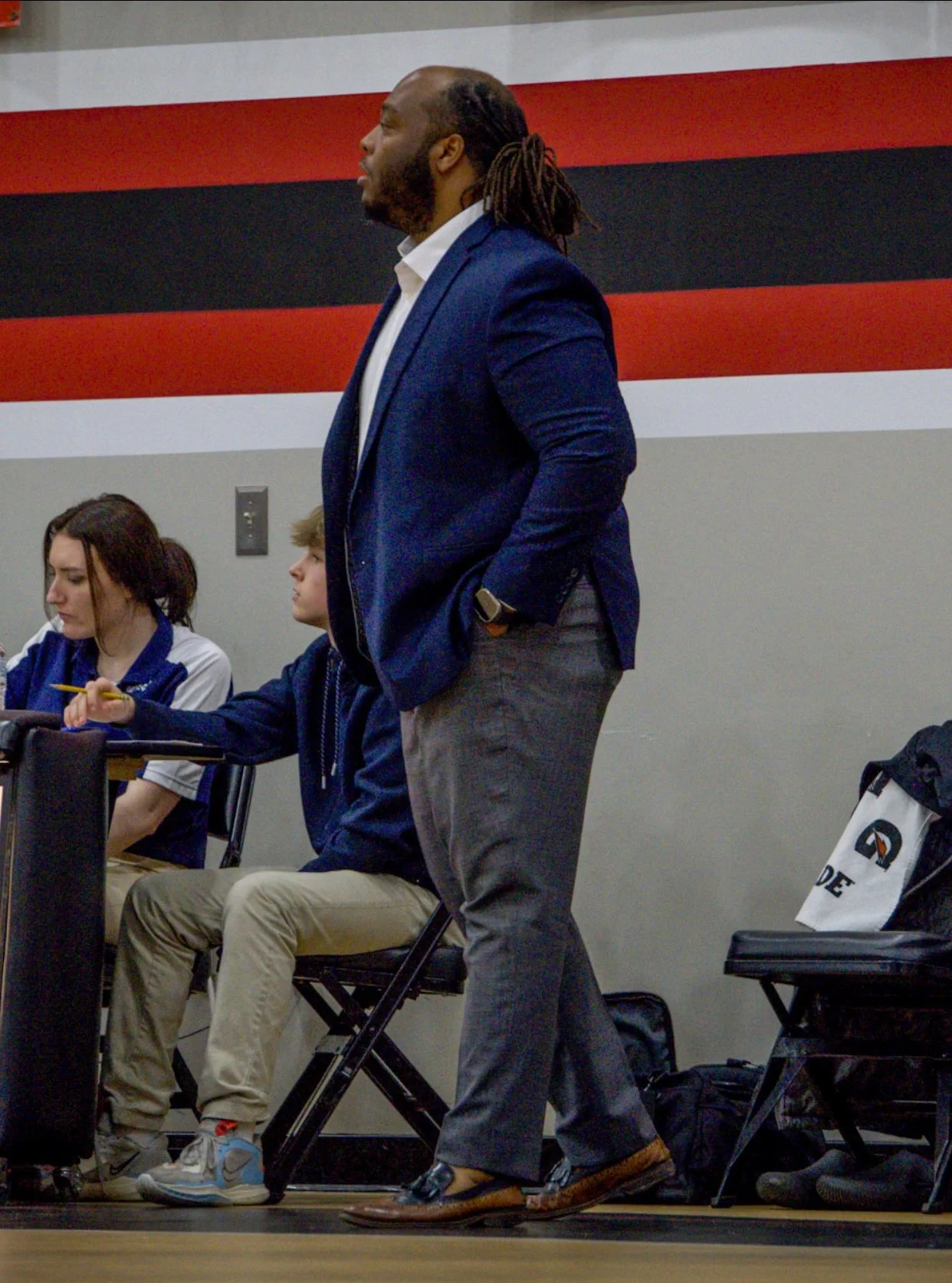 A man with dreadlocks and a beard, wearing a blue blazer and gray pants, stands with his hands in his pockets in a classroom or conference room. Several students are seated at desks, one is taking notes, and a backpack is on a chair nearby. The wall behind has red, black, white, and gray stripes.