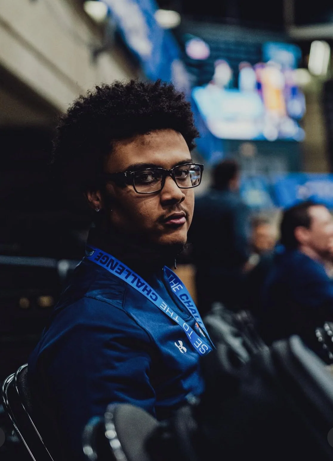 A young man with glasses and curly hair sitting in a stadium, wearing a blue jacket and a lanyard that says 'SEASON' and 'THE CHALLENGE'.