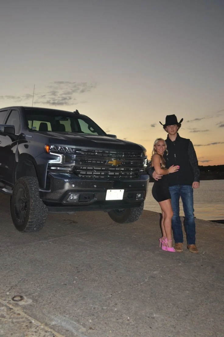 A young couple stands by a black Chevrolet SUV near a body of water at sunset. The woman is wearing a black dress and pink high heels; the man is dressed in dark jeans, a cowboy hat, and a western-style shirt.