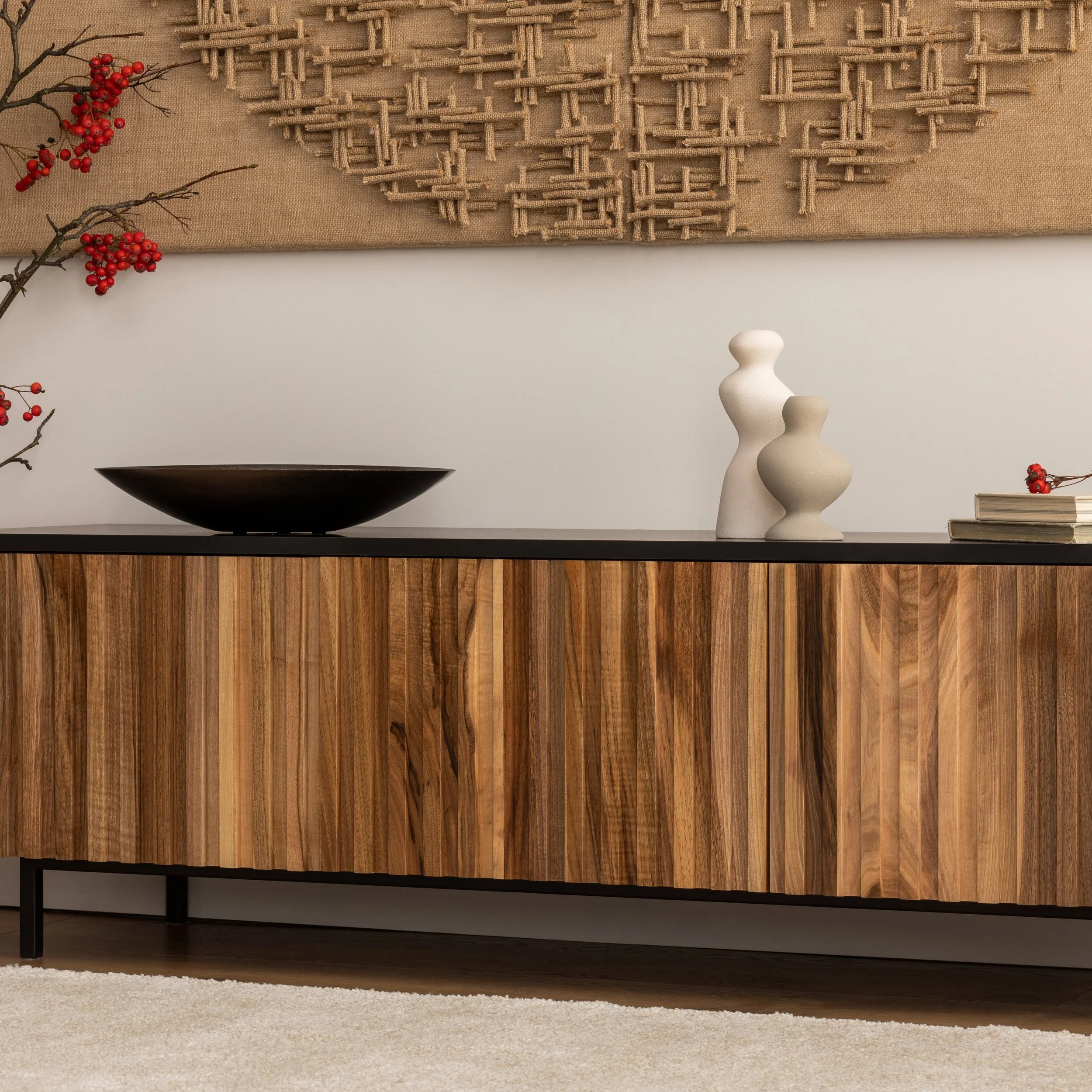 Une meuble en bois avec deux vases blancs et une coupe noire, décorée de branches de baies rouges, avec une œuvre d'art en bois suspendue au mur.