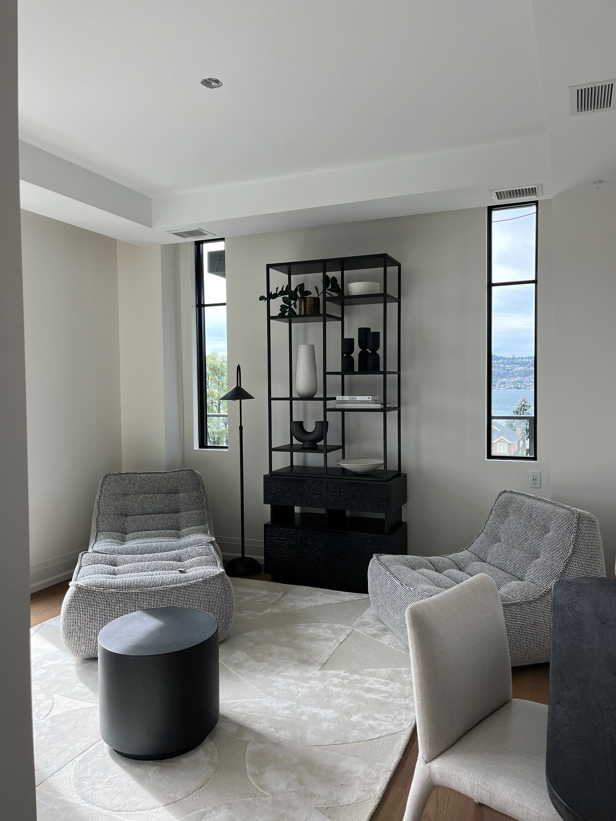 Modern living room with two gray armchairs, a black round side table, a black shelving unit with decorative vases and books, a black floor lamp, and windows showing a cityscape and water view.
