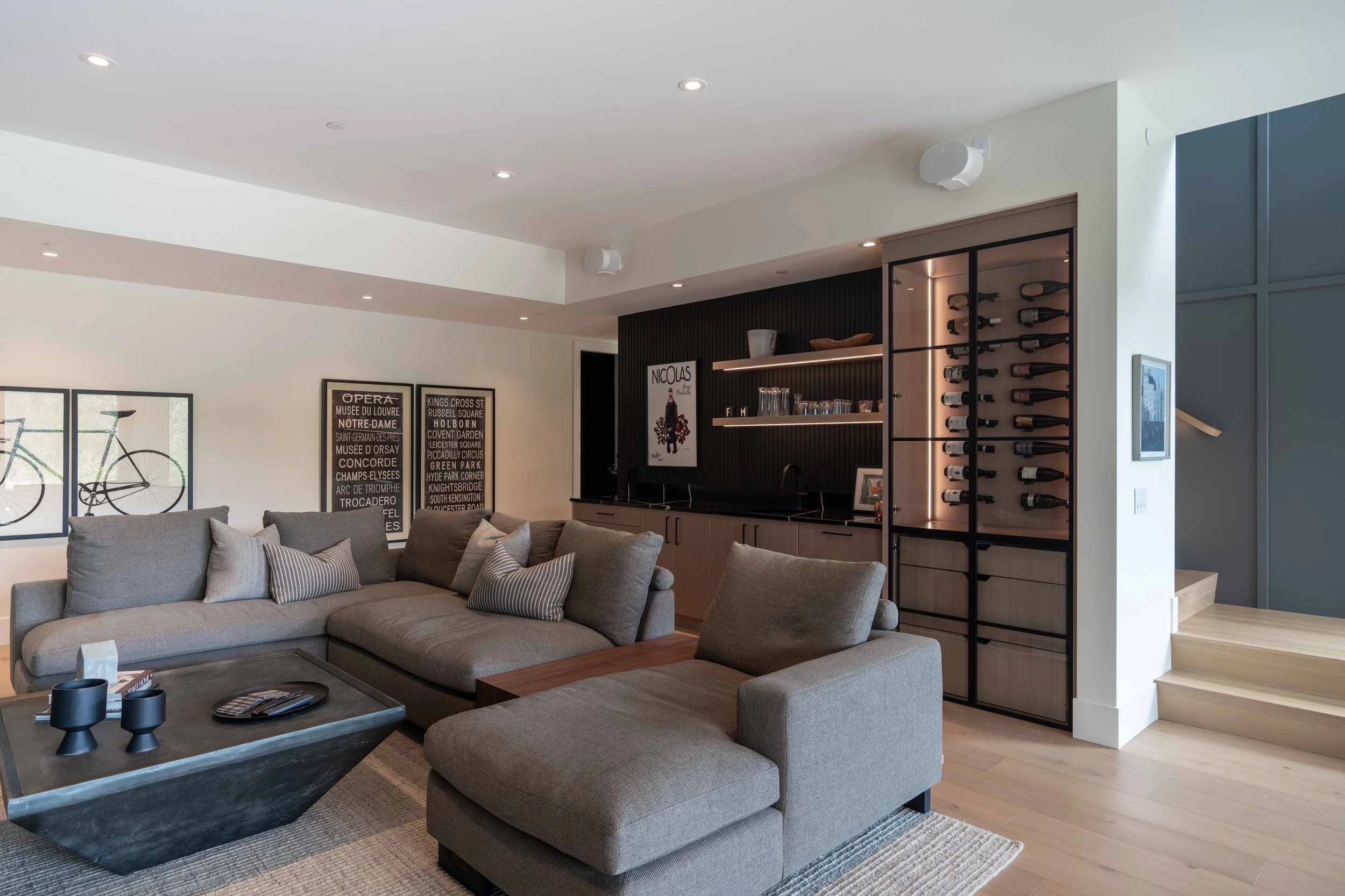 Modern living room with beige sectional sofa, striped pillows, black coffee table, wall art, bar area with shelves, wine rack, and steps to an upper level.