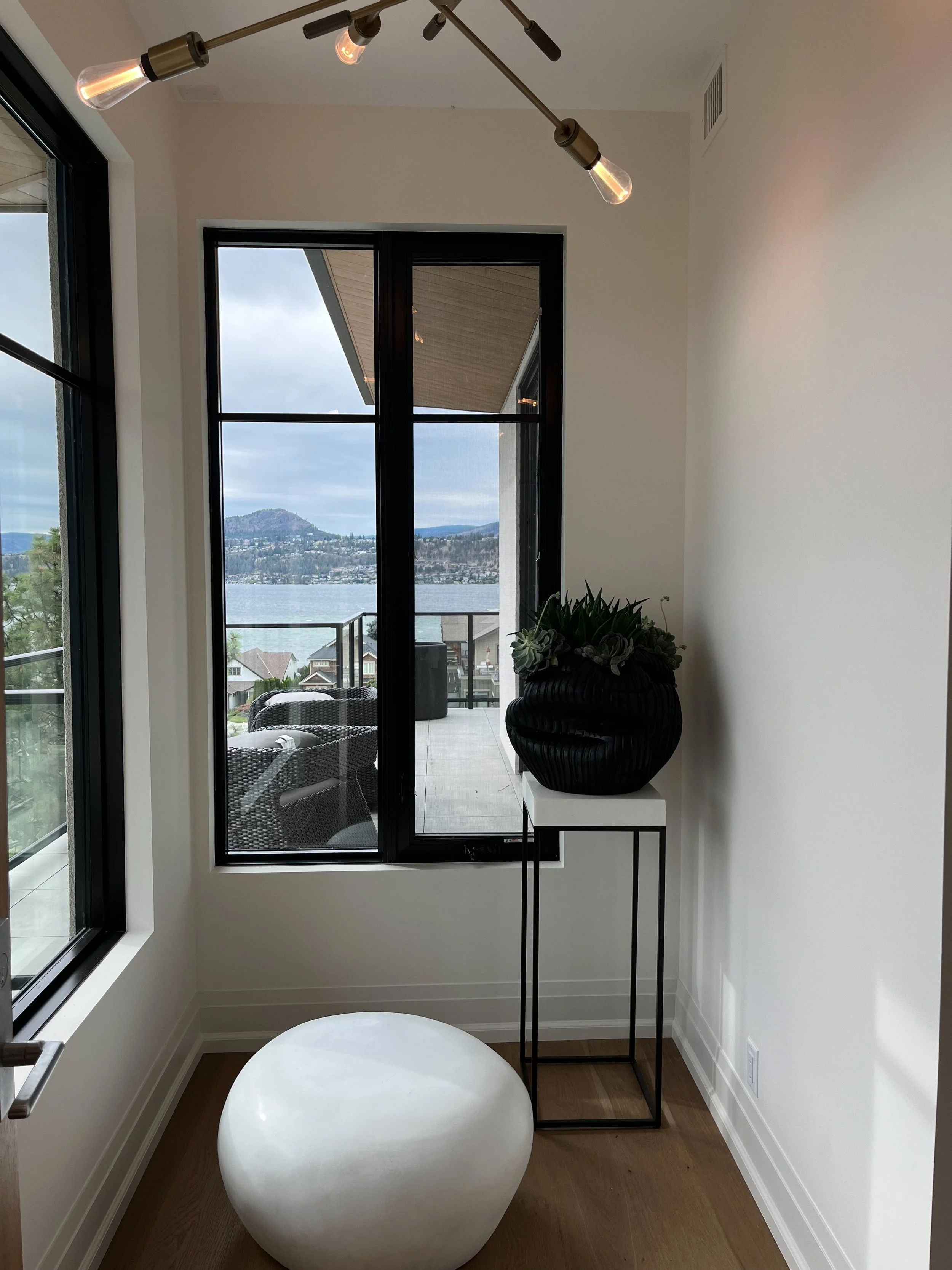 A corner of a room with large windows showing an outdoor balcony and a lake view. Inside, there is a modern white ottoman, a tall black vase with green plants on a stand, and a ceiling light fixture with exposed bulbs.
