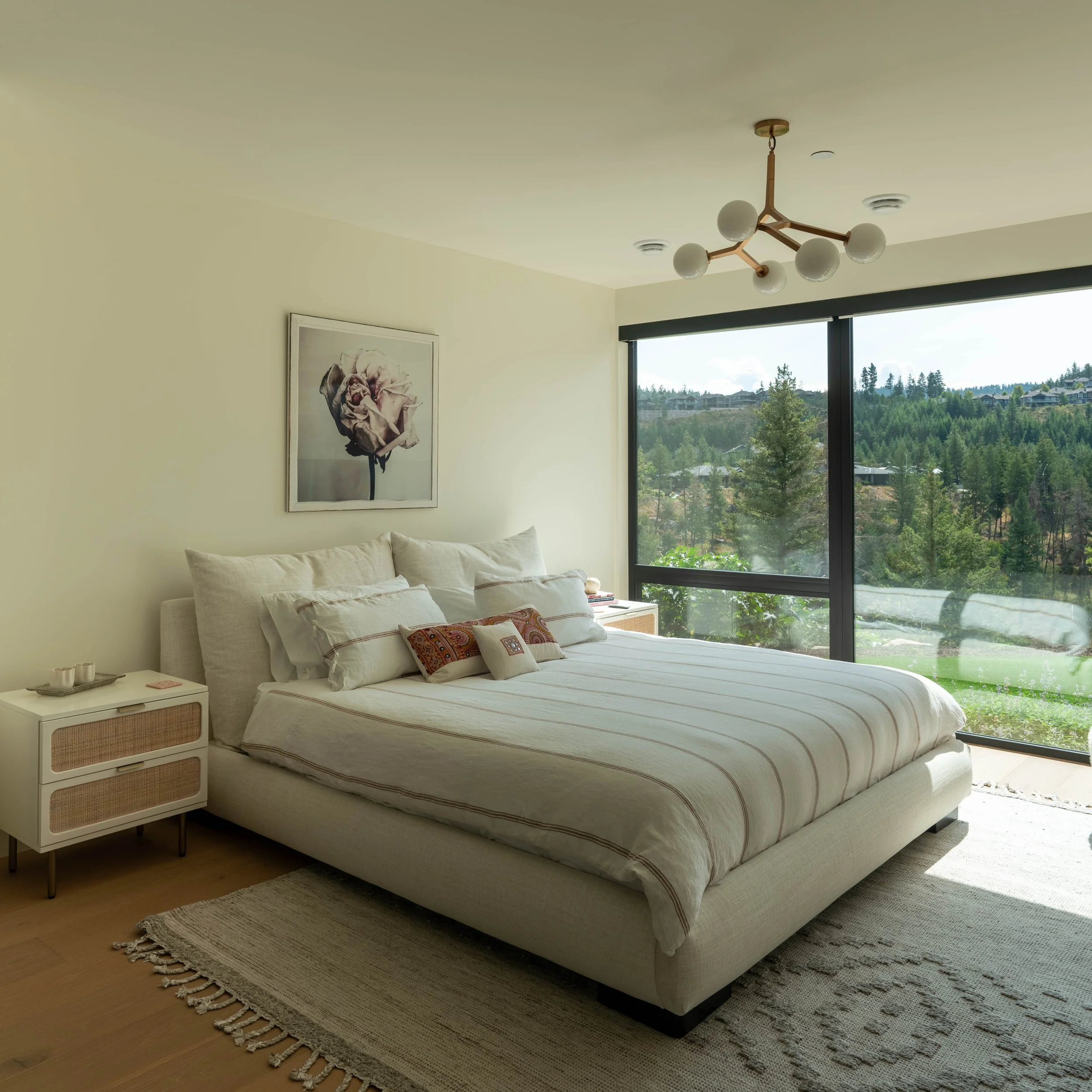A bedroom with a large bed, a nightstand, a framed flower picture, a window showing a green landscape, and a modern ceiling light.