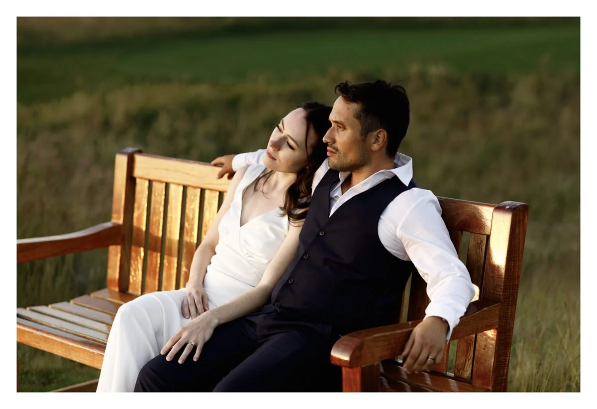 Wedding couple sitting together on a wooden bench during sunset at Kinkell Byre, overlooking St Andrews Bay, with the bride resting her head on the groom's shoulder and both relaxed and content