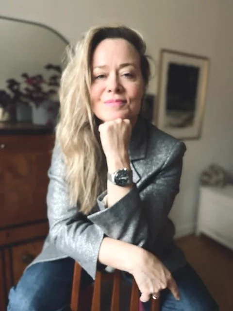 Woman with long blonde hair, sitting with chin resting on fist, wearing a silver blazer and jeans, in a room with a dresser, mirror, and framed picture in the background.