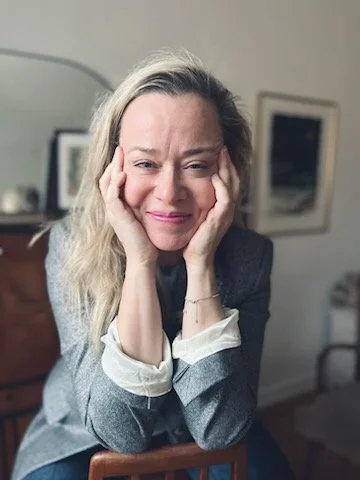 A woman with blonde hair, smiling with her hands on her cheeks, seated in a room with artwork and furniture.